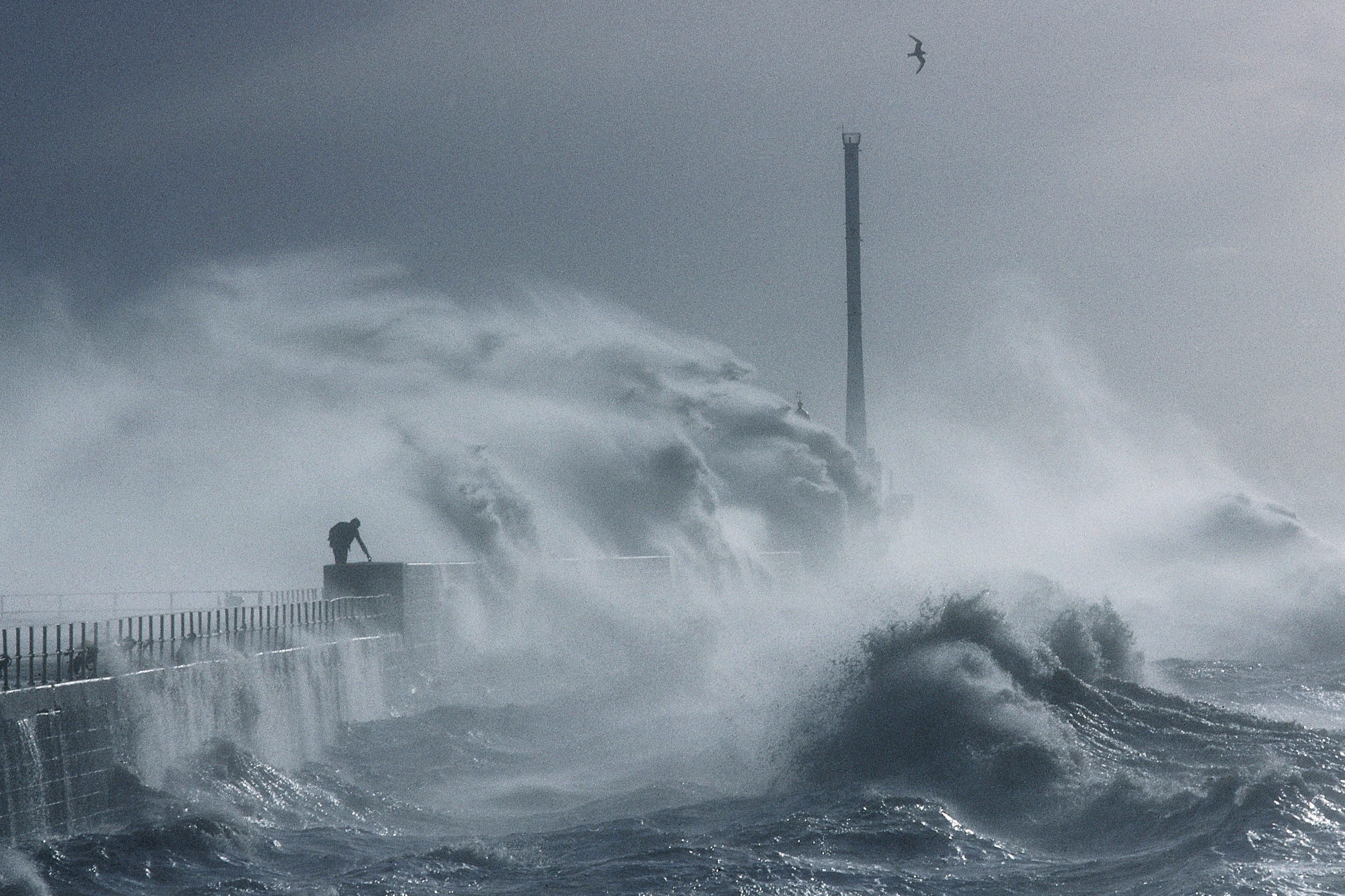 Photo of a pier with large crashing waves and mist, a person on the pier and a bird flying above in the stormy sky.