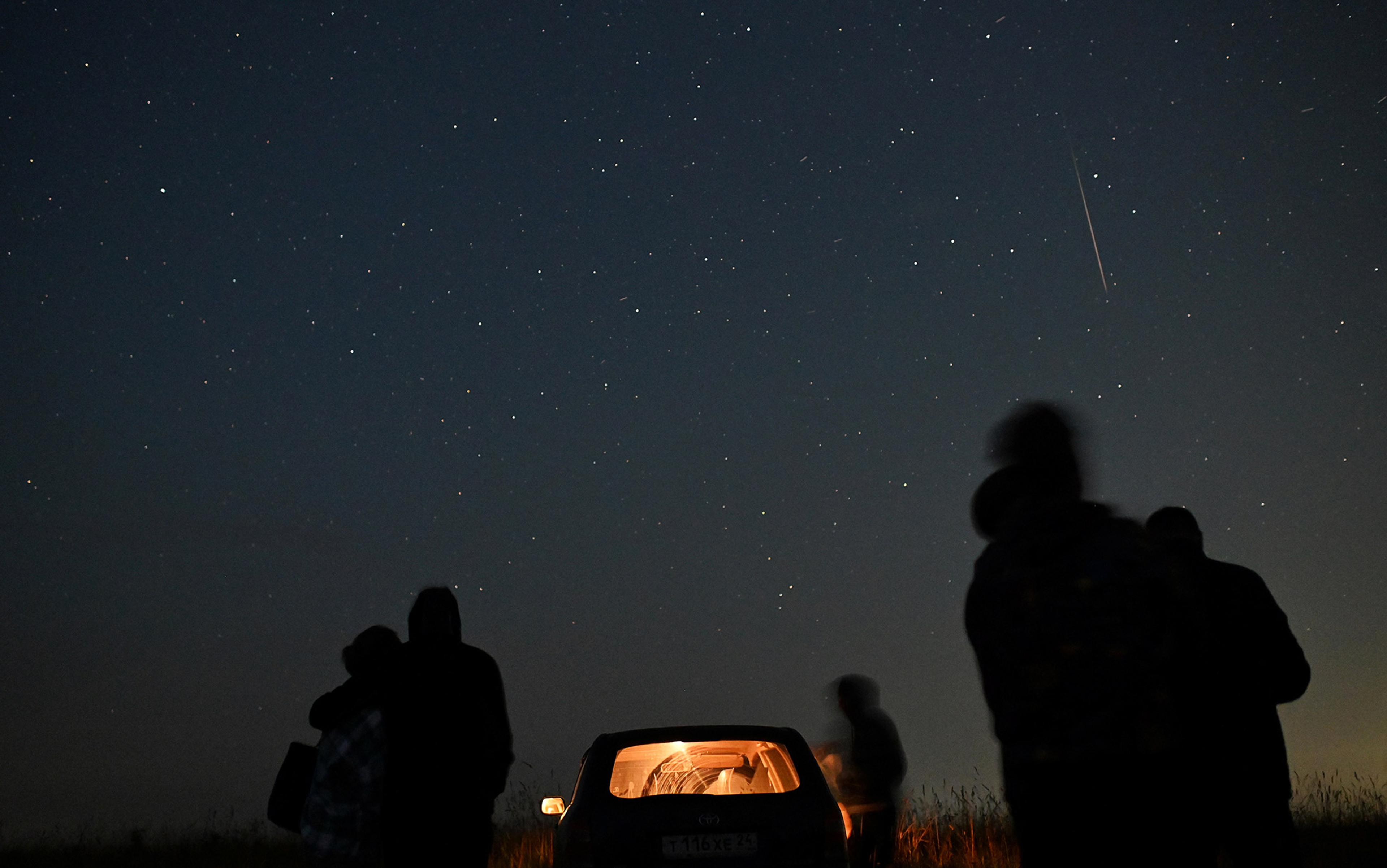Silhouetted figures stand beside a car looking up at a starry night sky