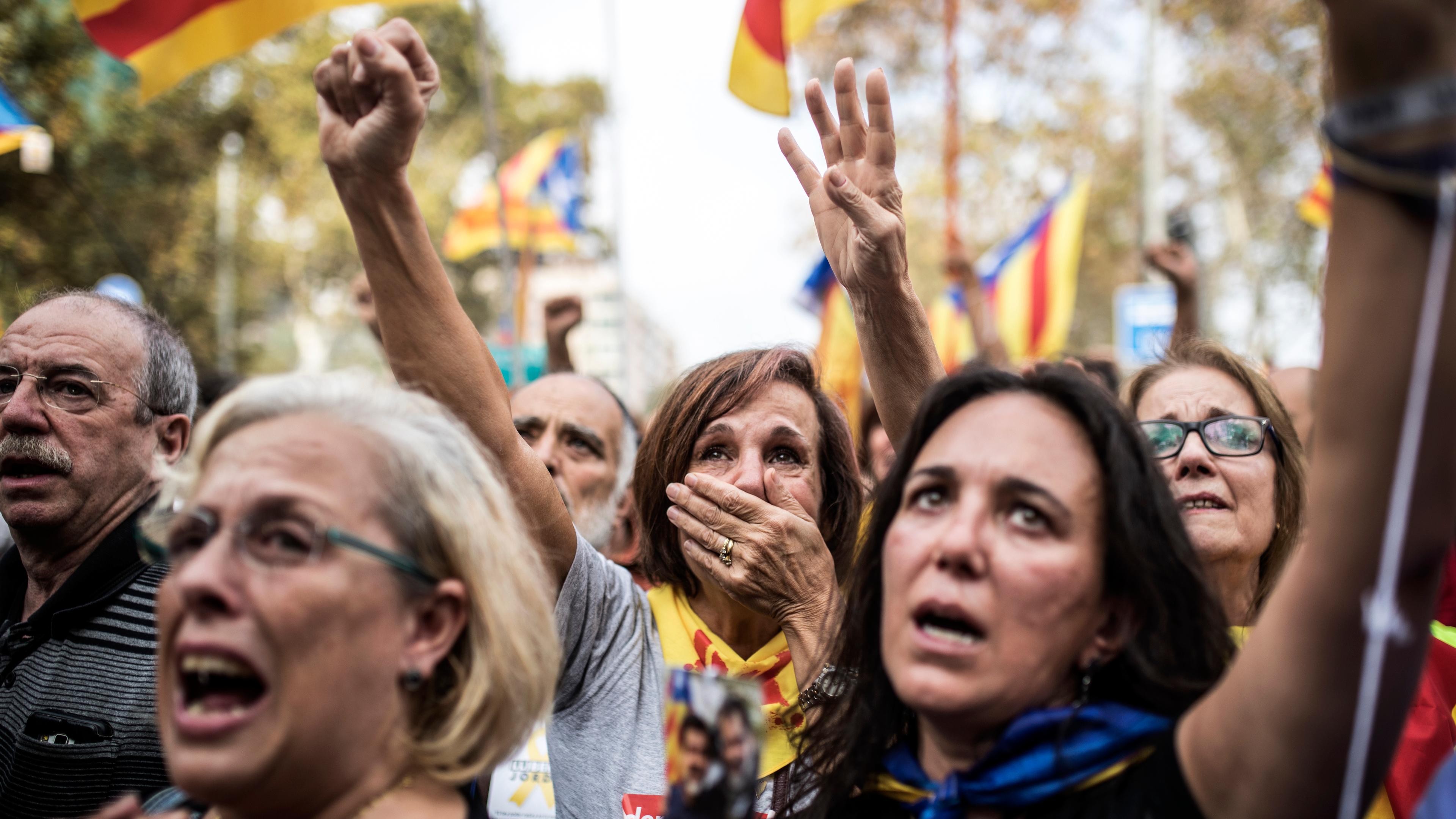 Photo of emotional protesters raising hands and holding Catalan flags in a crowd during a demonstration.