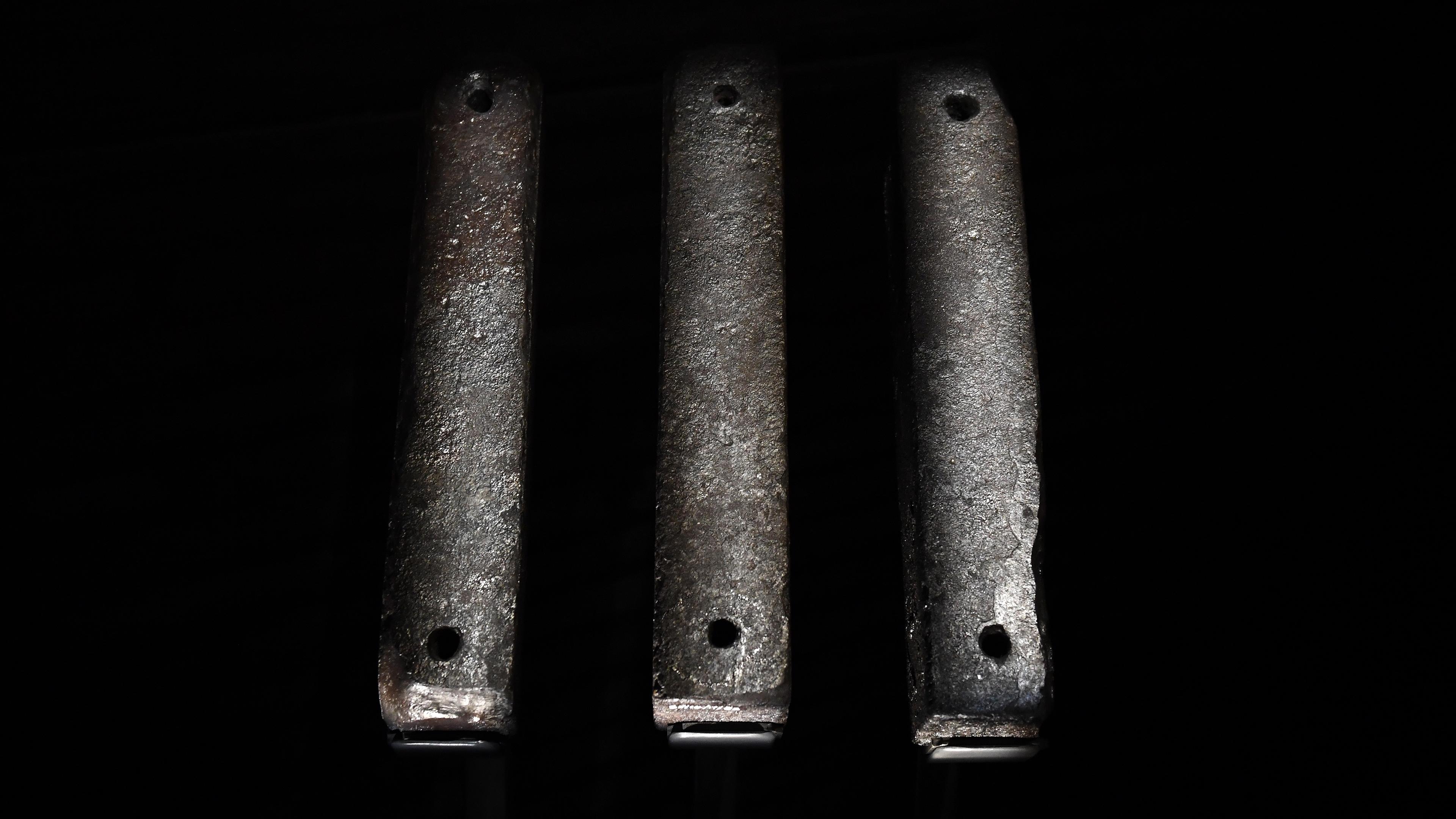 Photo of three aged metal bars with small holes, standing vertically against a dark background.