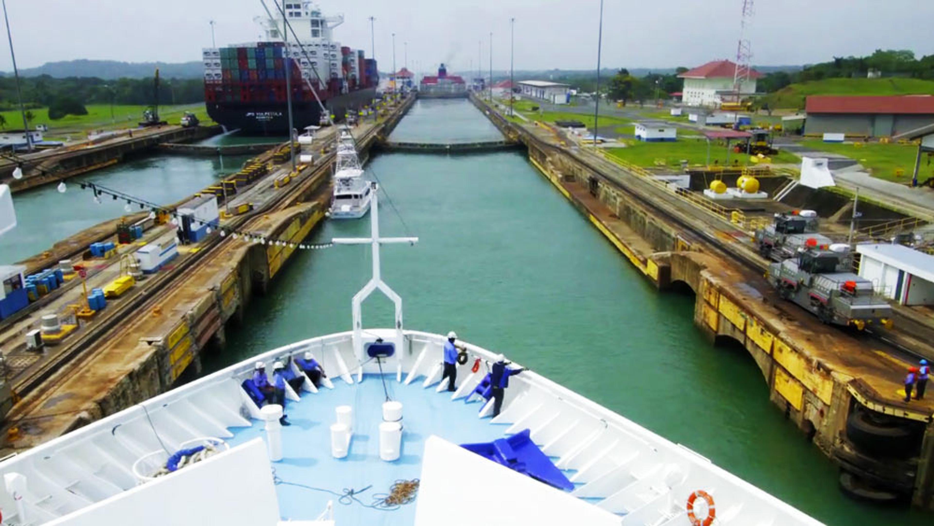 A ship’s bow entering a lock in the Panama Canal with a large container ship ahead and industrial infrastructure on both sides.
