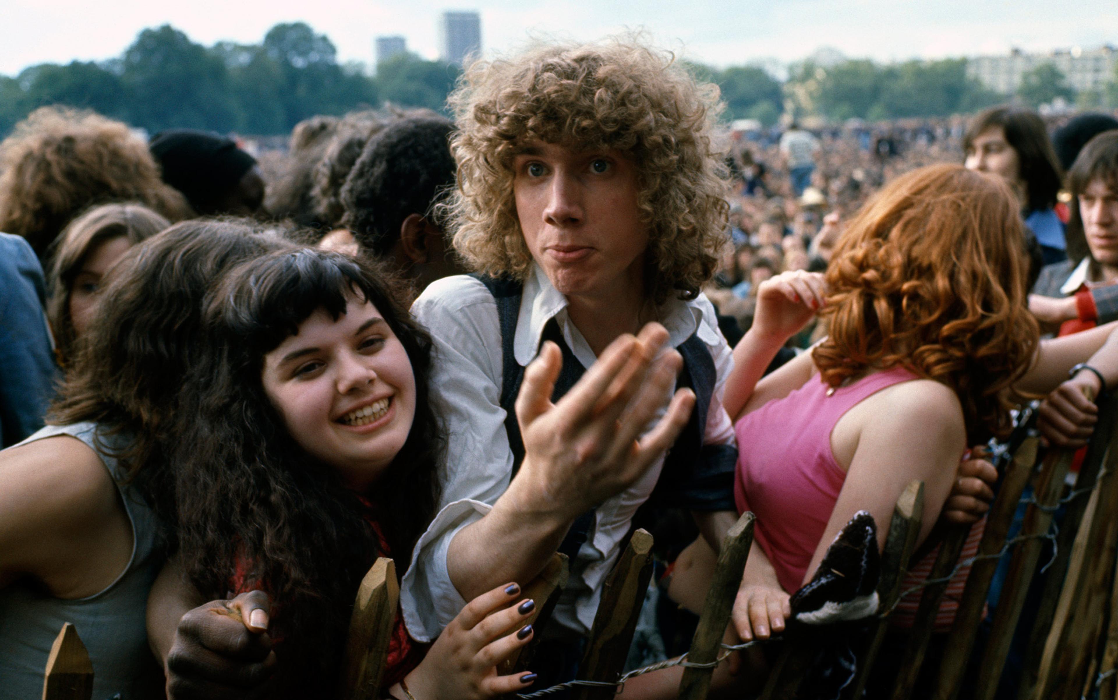 A young crowd at an outdoor event with people smiling and leaning on a wooden barrier, trees and buildings in the background.