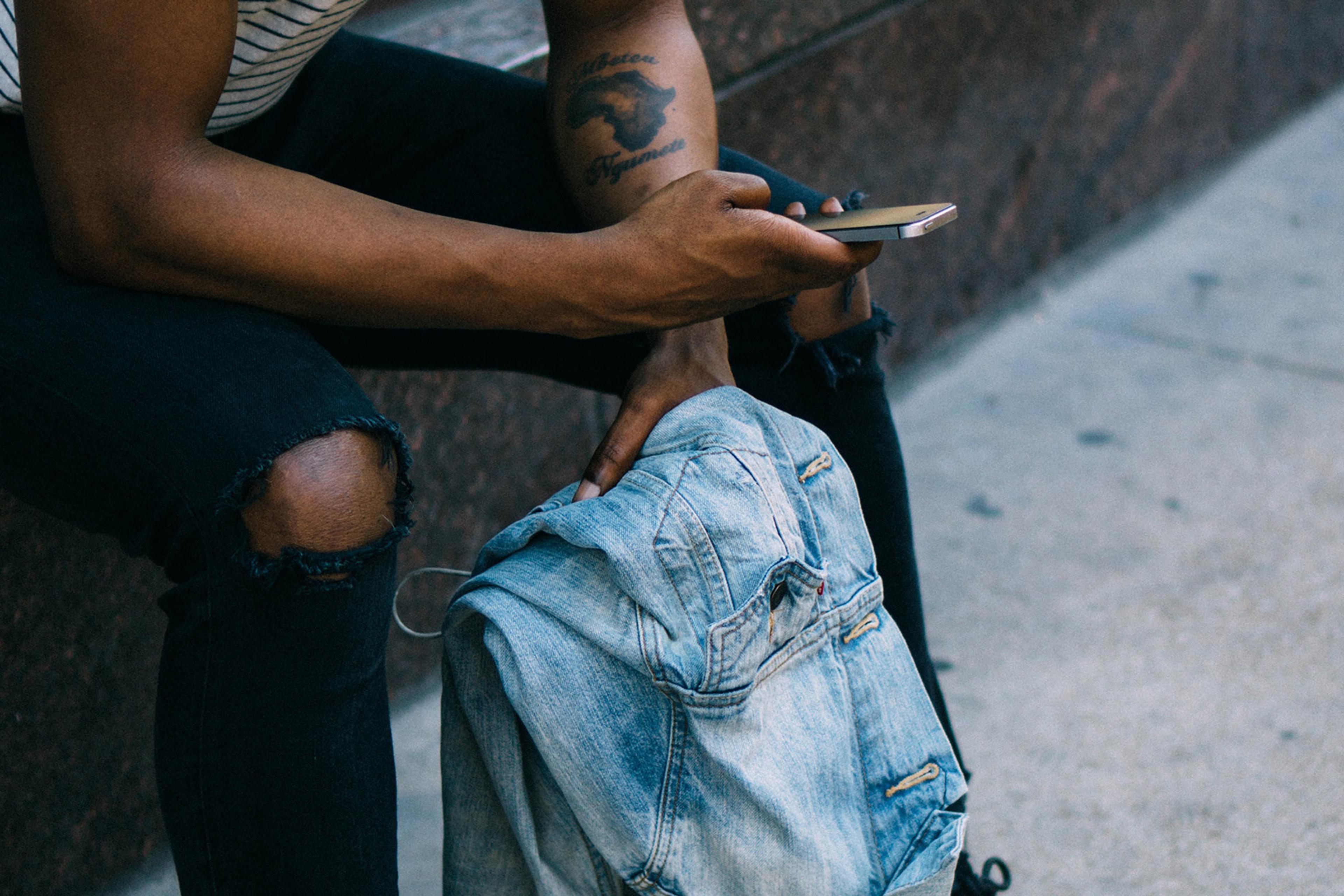 A person with tattoos and wearing ripped jeans sits on a bench using a phone with one hand and holding a denim jacket with the other.