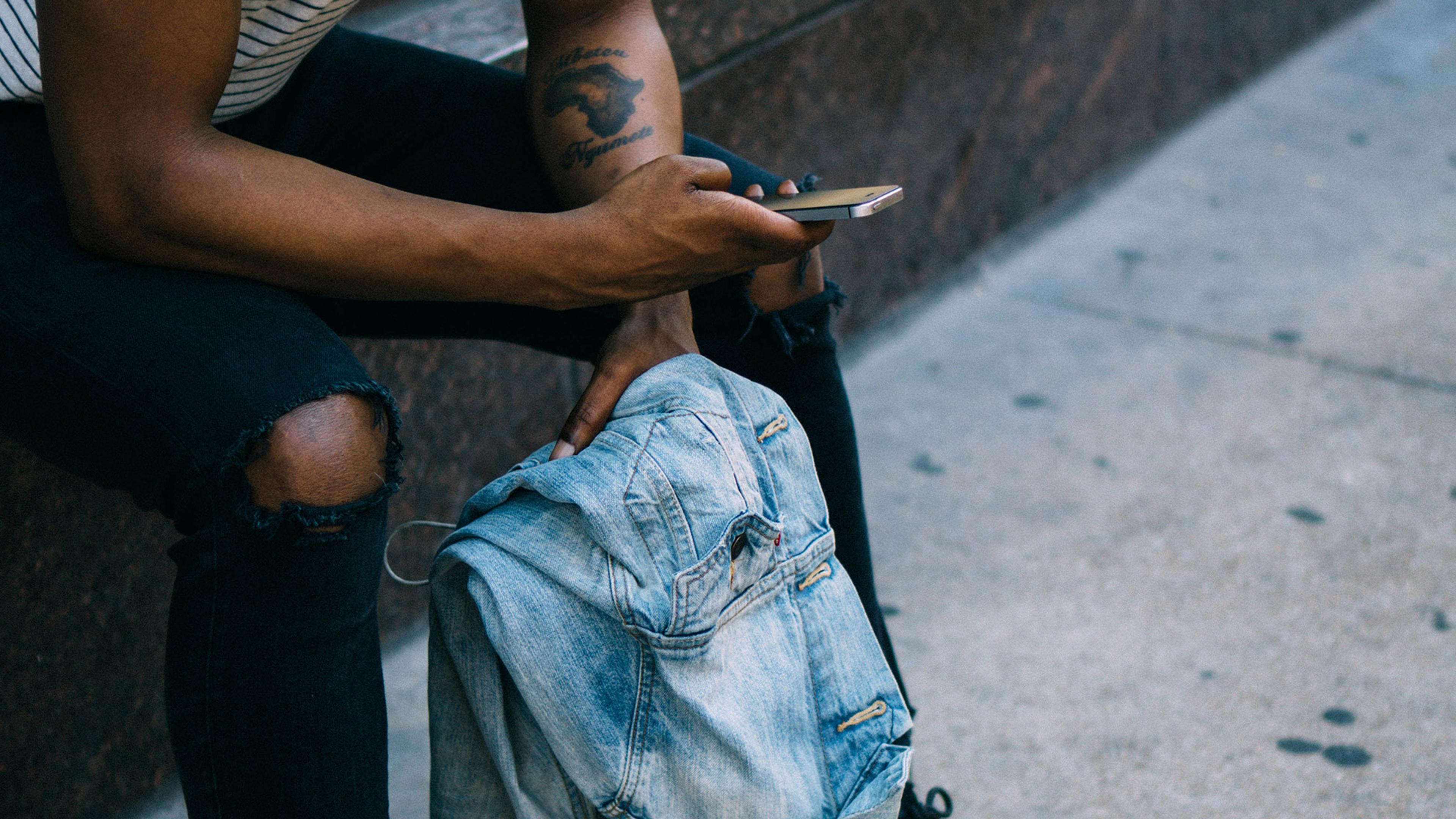 A person with tattoos and wearing ripped jeans sits on a bench using a phone with one hand and holding a denim jacket with the other.