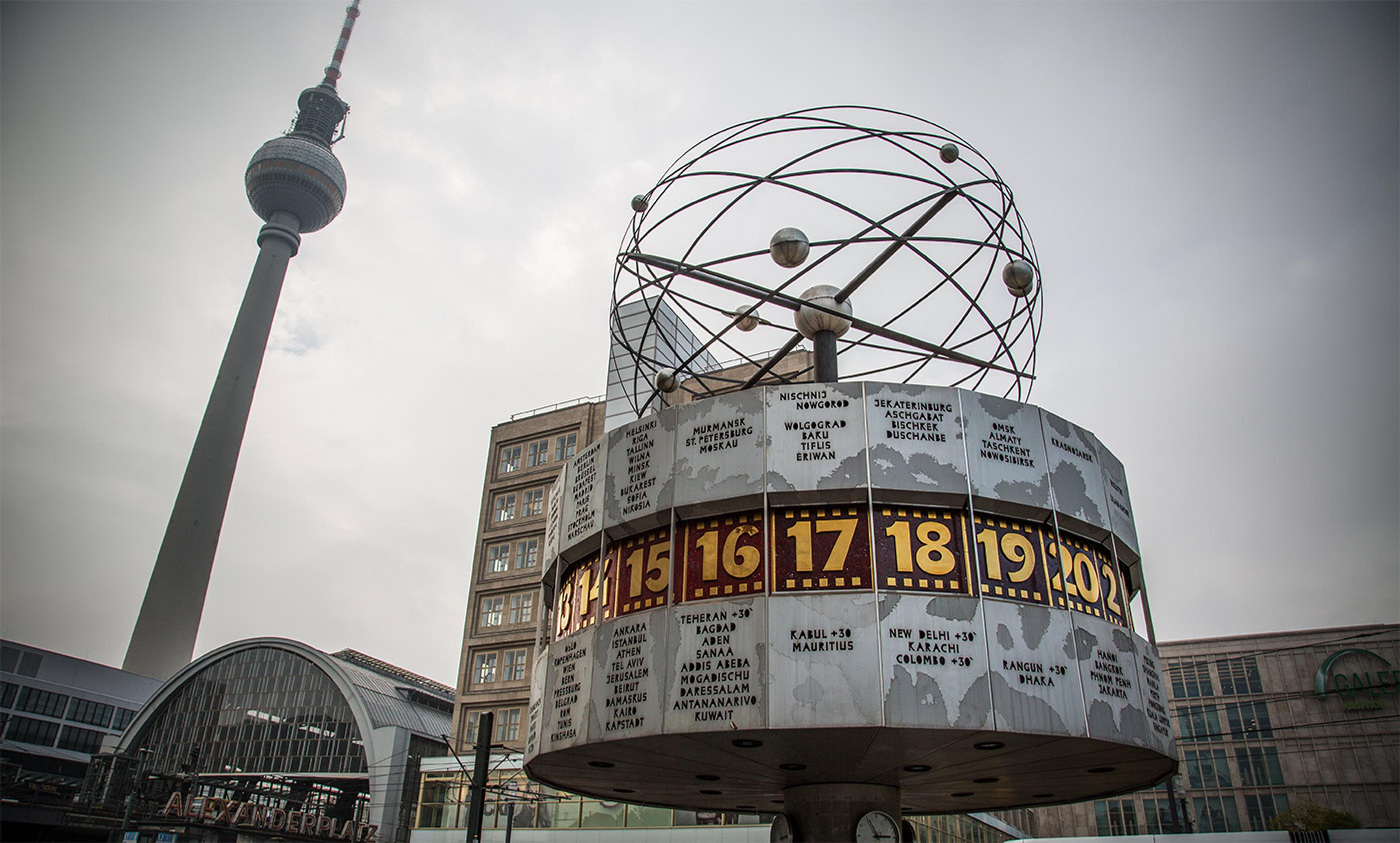 The World Clock and Fernsehturm at Alexanderplatz Berlin featuring city names and times with surrounding buildings and a cloudy sky.