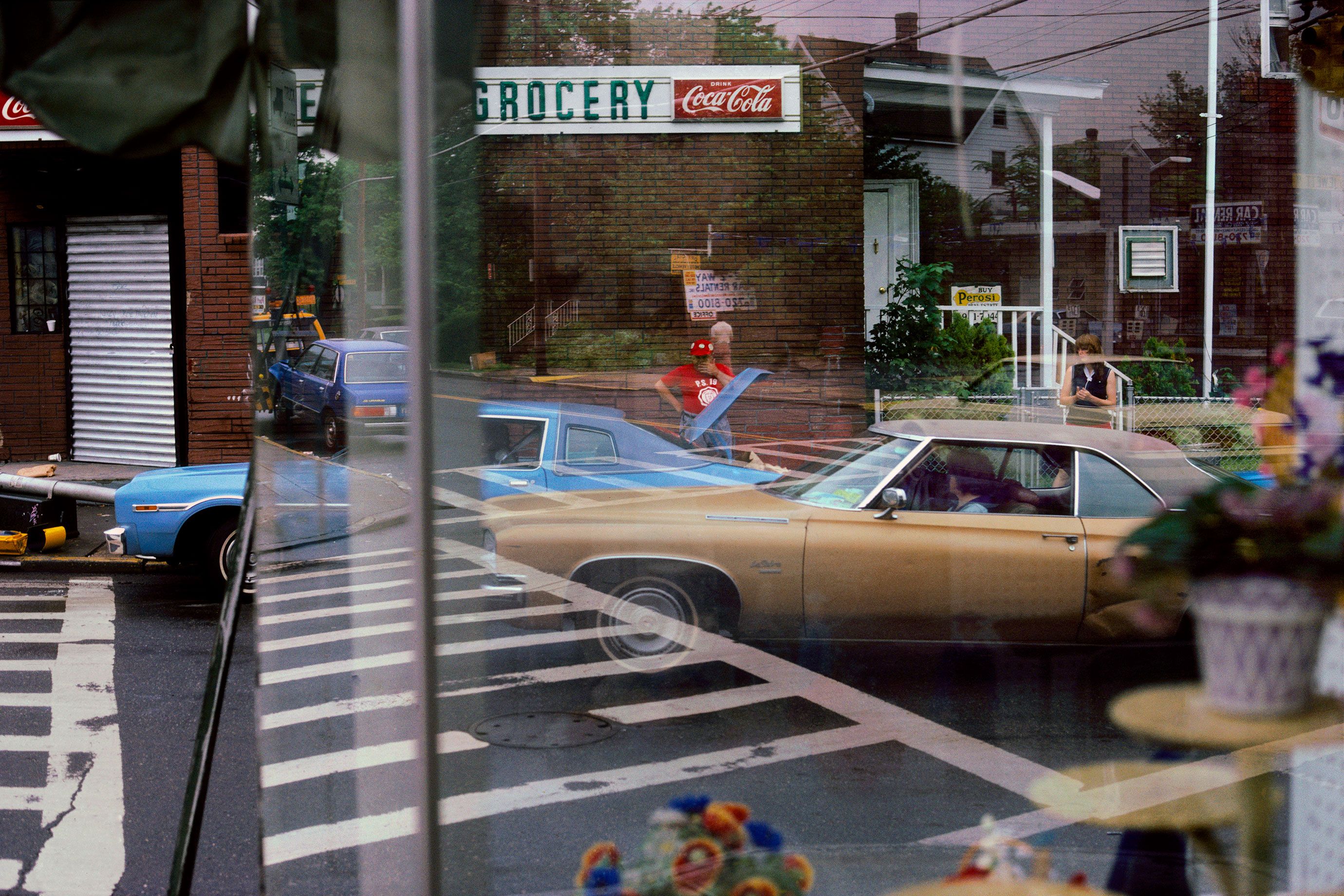 A city street with reflections on a window showing cars, people and a grocery sign with a Coca-Cola advertisement.