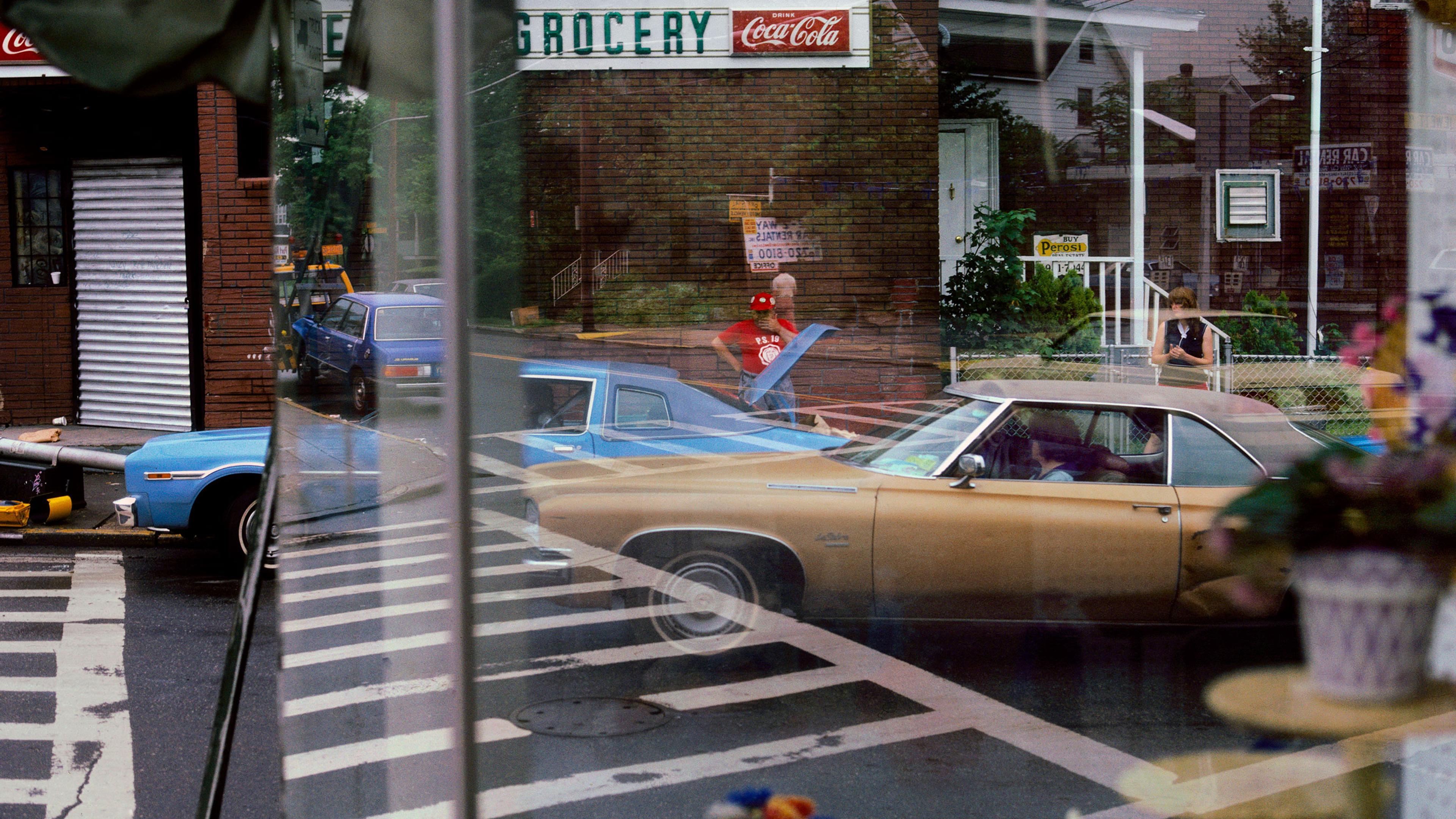 A city street with reflections on a window showing cars, people and a grocery sign with a Coca-Cola advertisement.