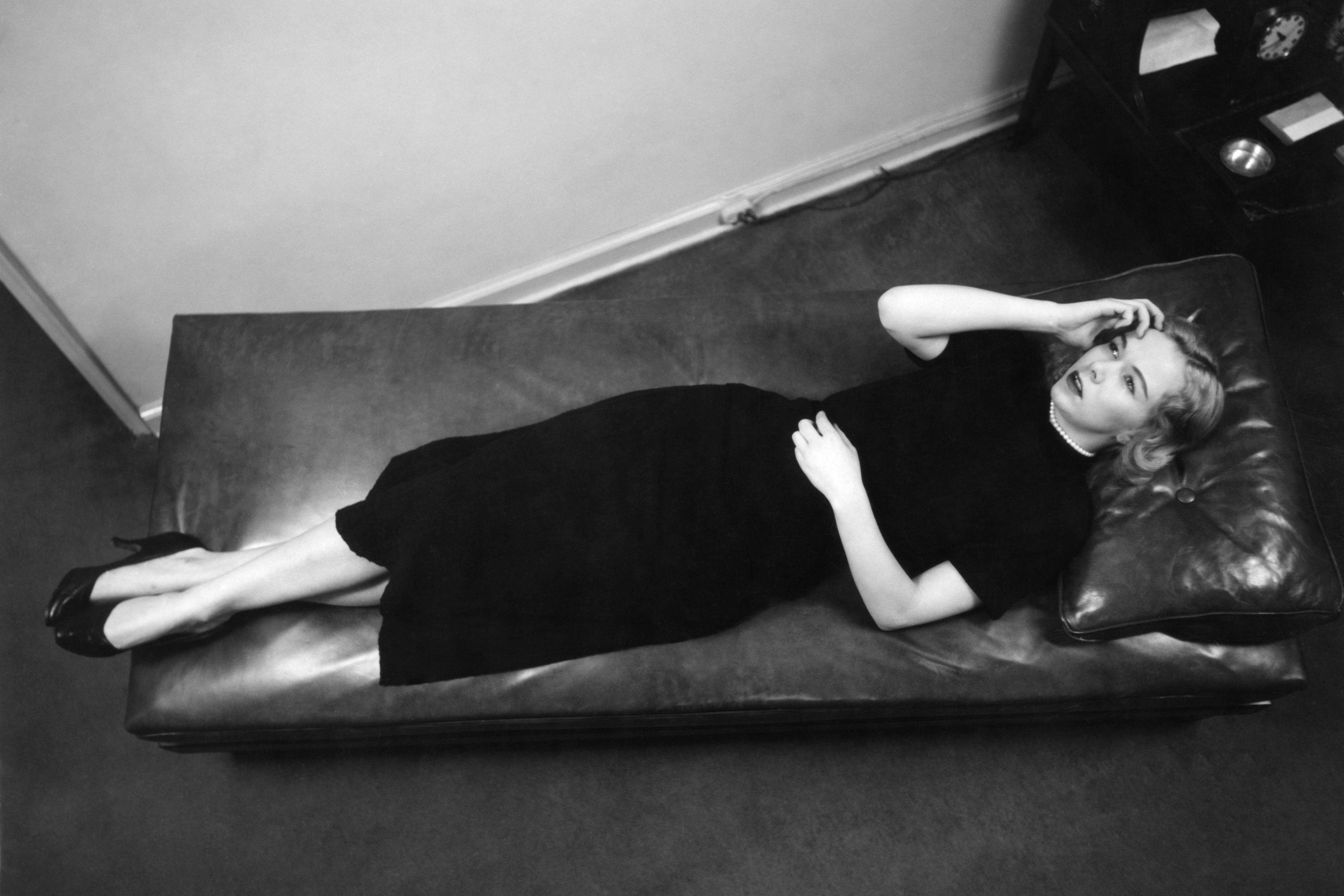 Black and white photo of a woman in a dark dress lying on a leather couch, looking upward with one hand on her face.