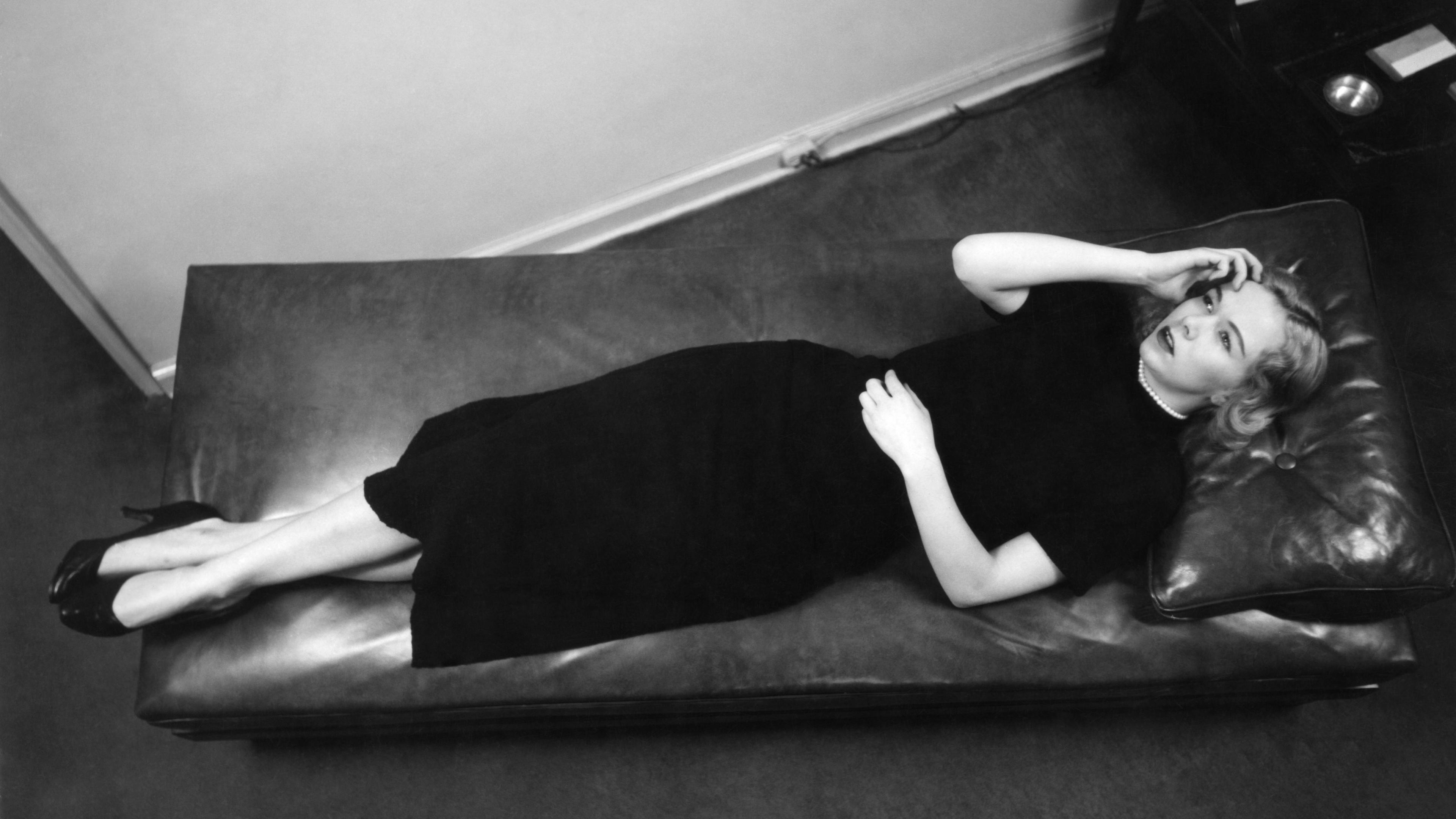 Black and white photo of a woman in a dark dress lying on a leather couch, looking upward with one hand on her face.
