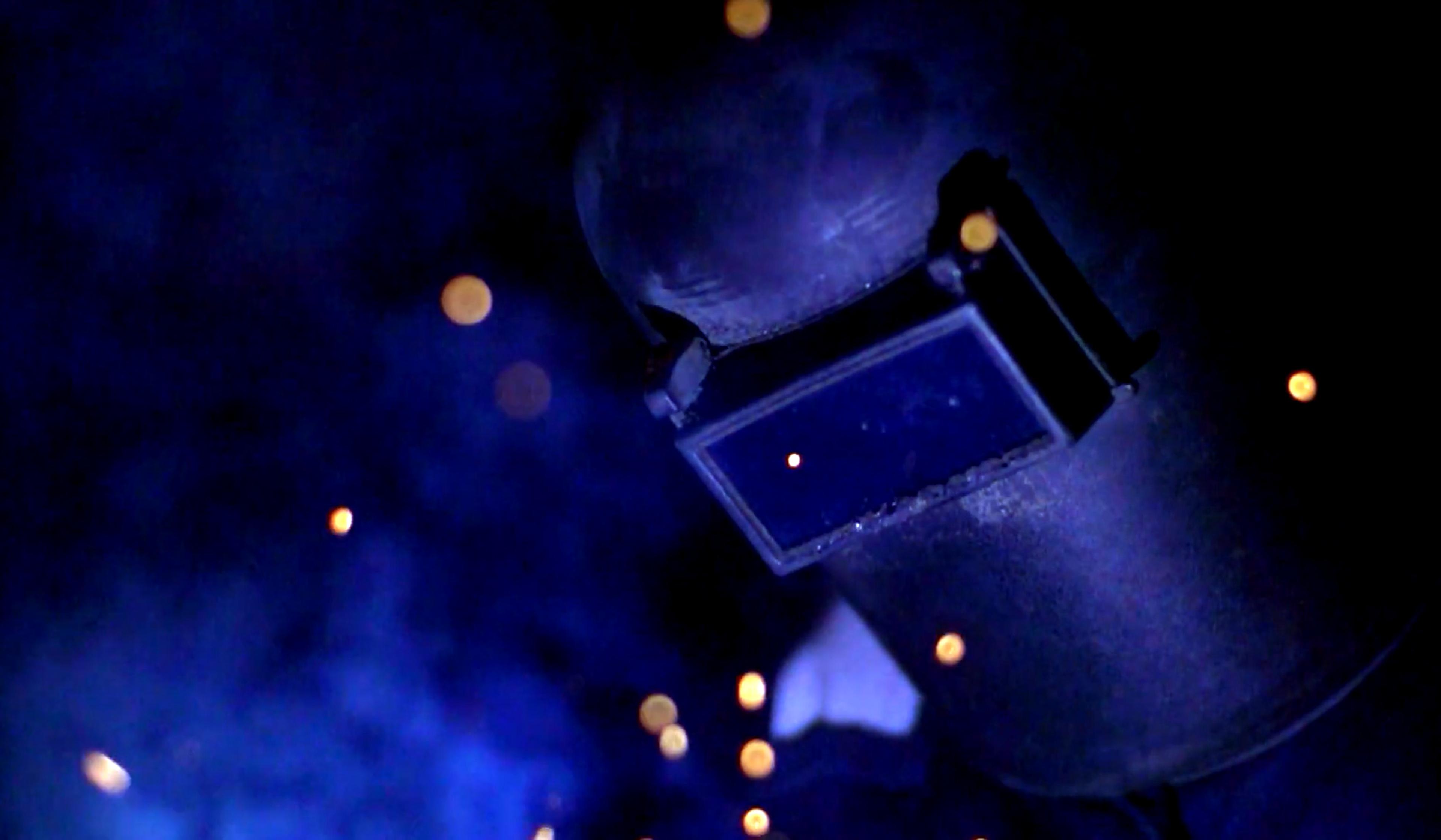 A welder wearing a protective mask surrounded by sparks and blue light. The scene is dimly lit, highlighting the sparks.