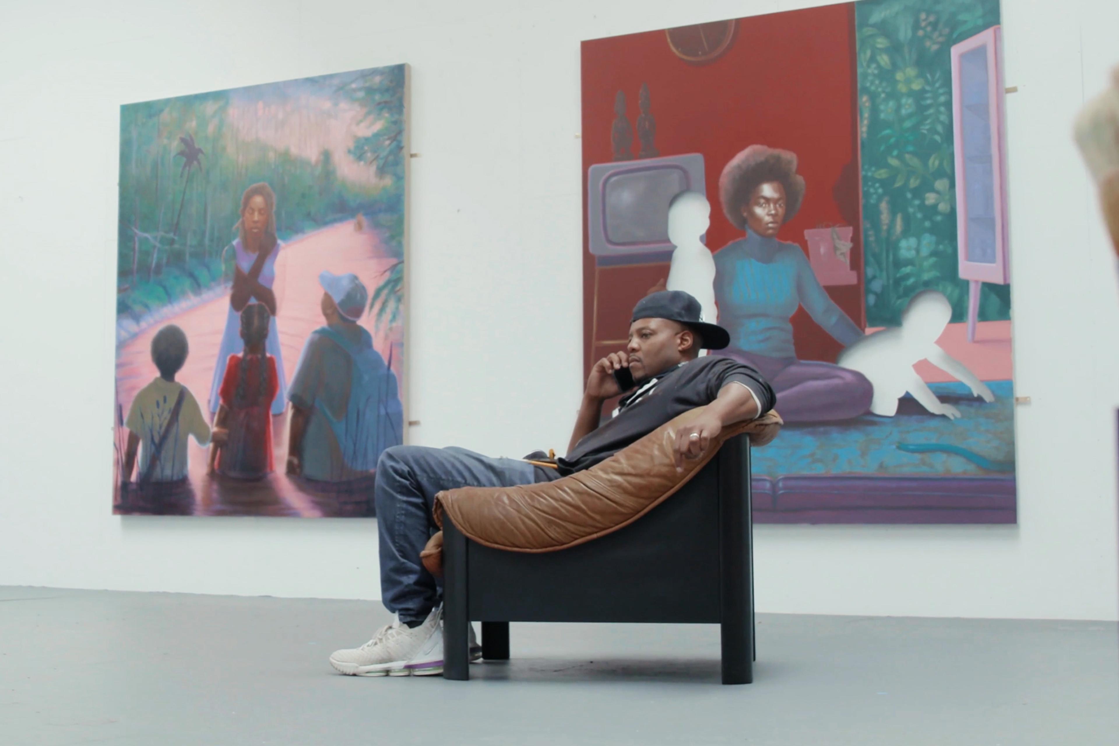 A person sits in a chair on the phone in an art gallery, with two large paintings in the background and paintbrushes visible in the foreground.