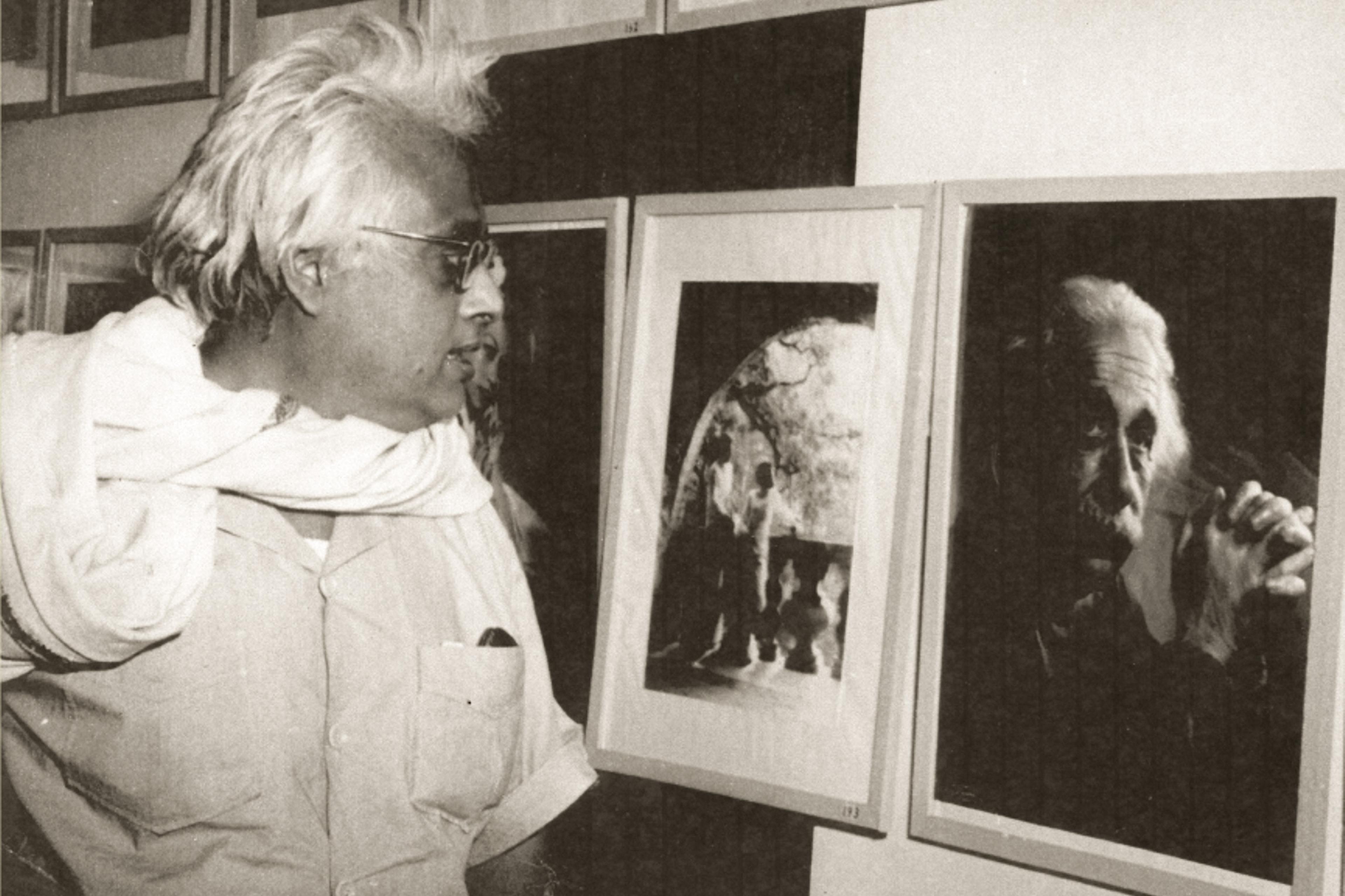 Black-and-white photo of a man viewing framed artworks on a wall in an exhibition setting.