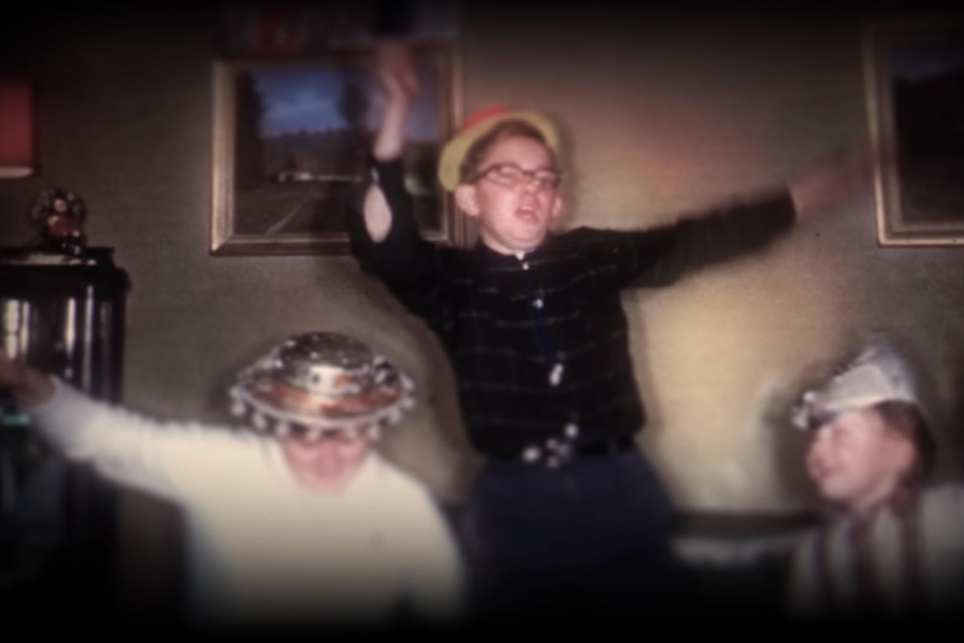 Vintage photo of three children dancing in hats in a living room with framed paintings on the wall.