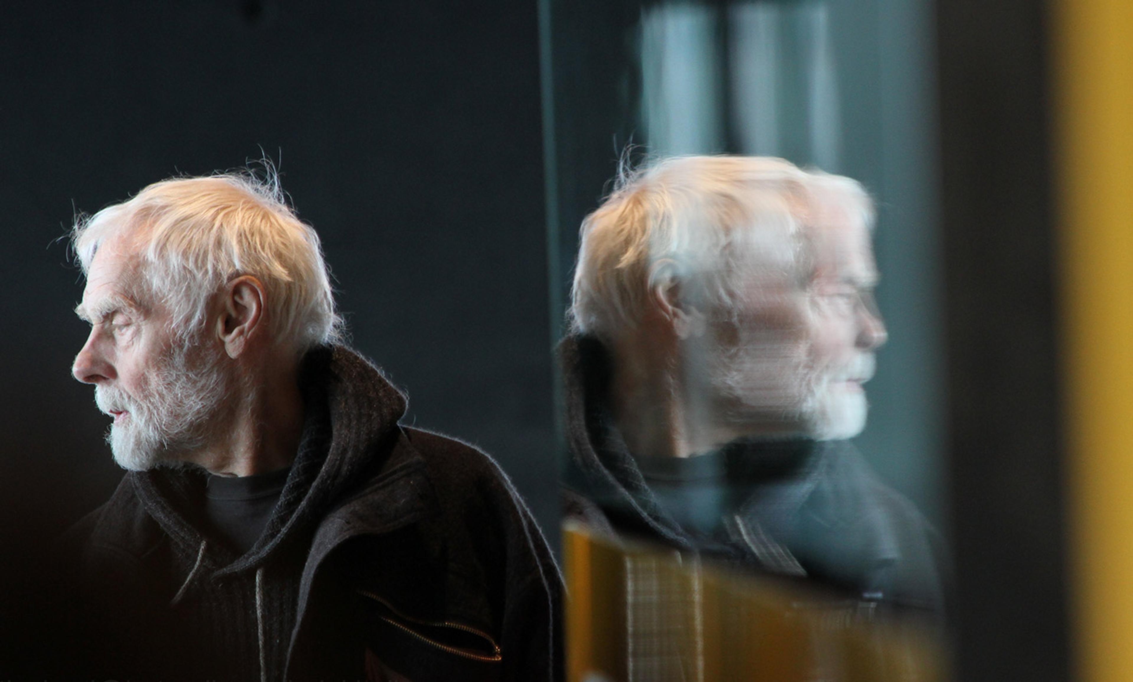 An elderly man with white hair and a beard looking to his left, with his reflection visible on a glass surface beside him