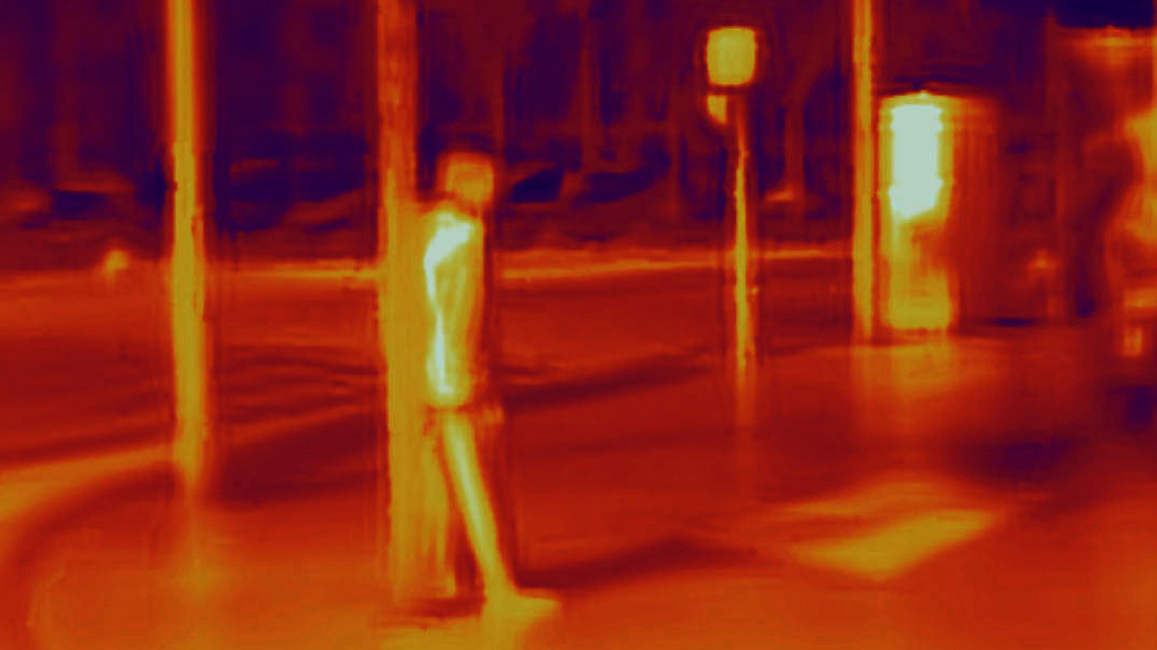Thermal image of a street scene with a person standing by a pole and bright signage in the background.