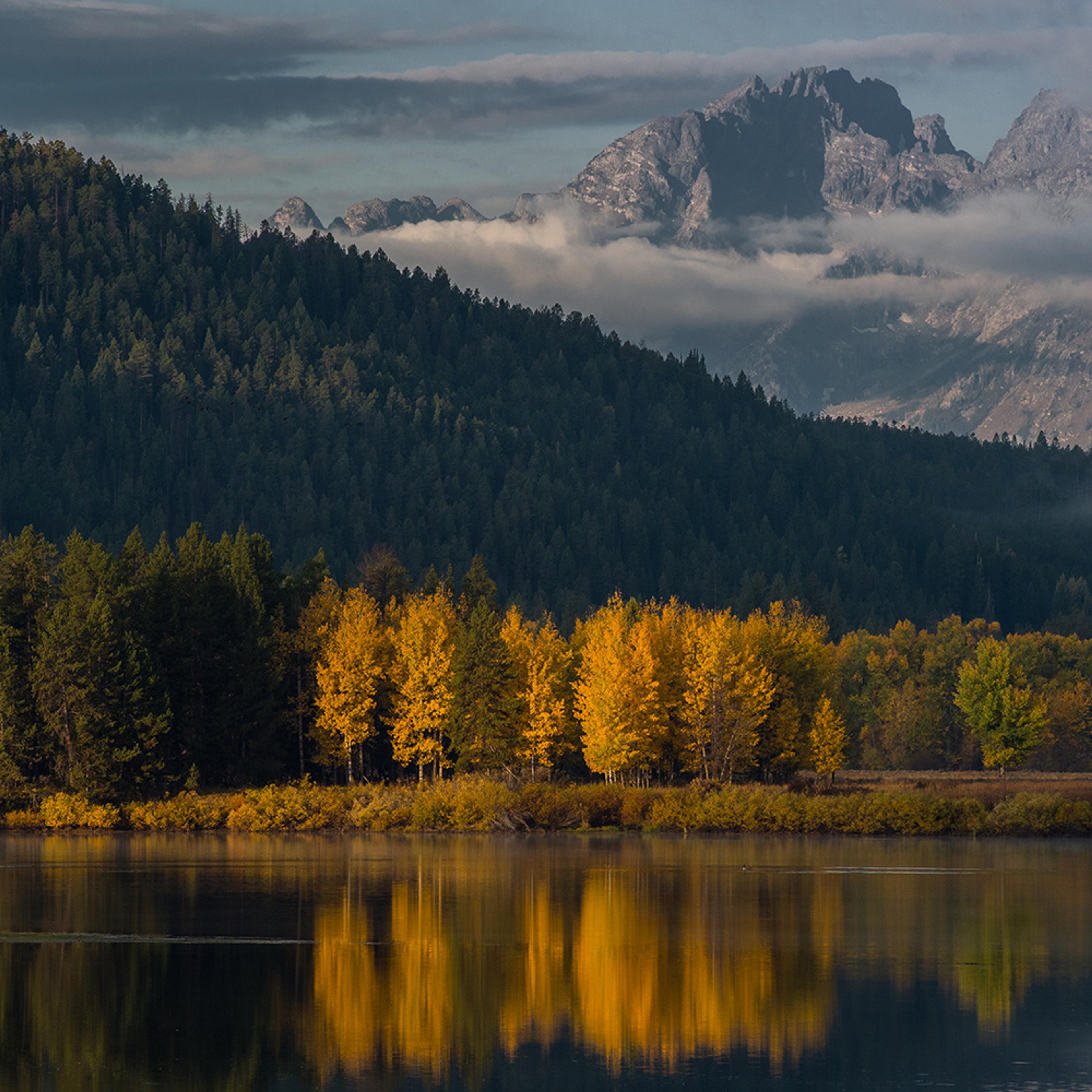 A lake with reflections of autumn trees, a dense forest beyond, mountains in the distance, and clouds overhead.