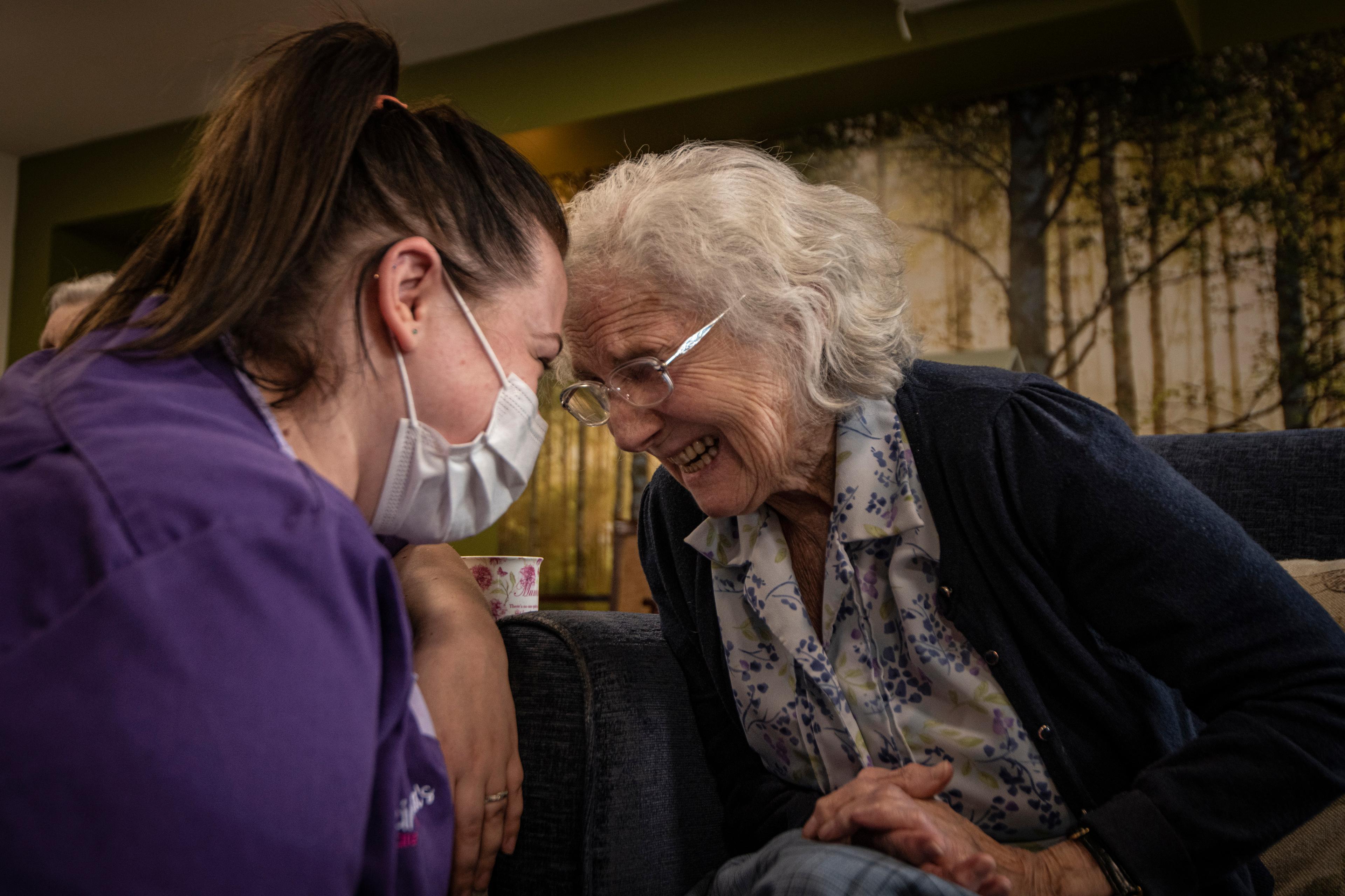 Photo of a young caregiver in a mask and an elderly woman smiling warmly, leaning towards each other in a cosy setting.
