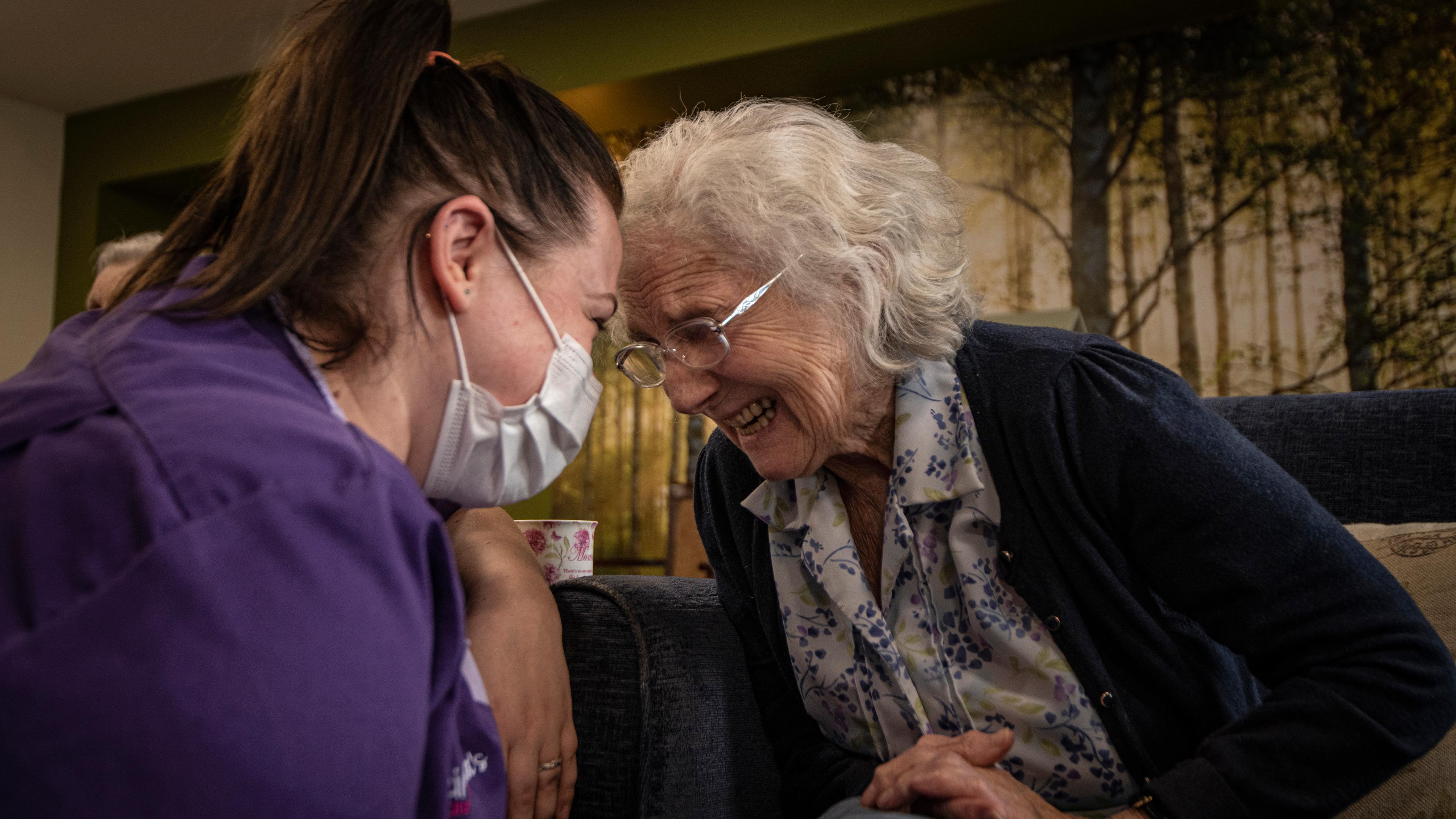 Photo of a young caregiver in a mask and an elderly woman smiling warmly, leaning towards each other in a cosy setting.