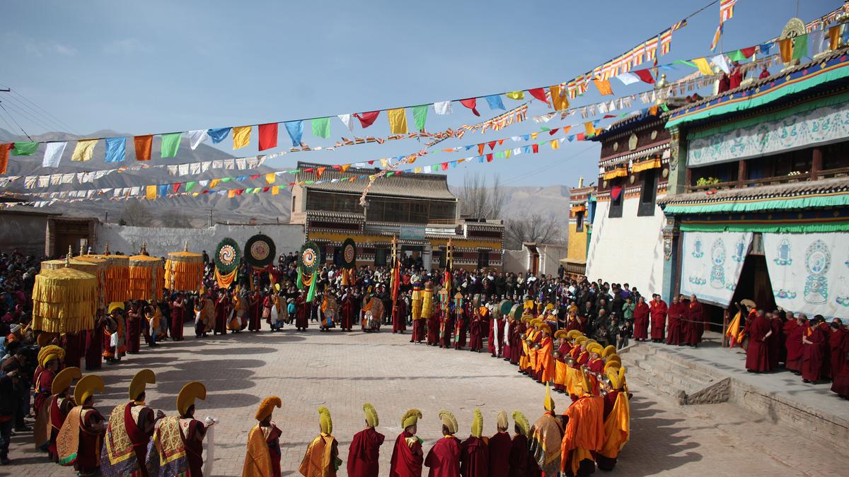 Monks in colourful robes and hats, forming a circle during an outdoor festival with flags and onlookers in Tibet.