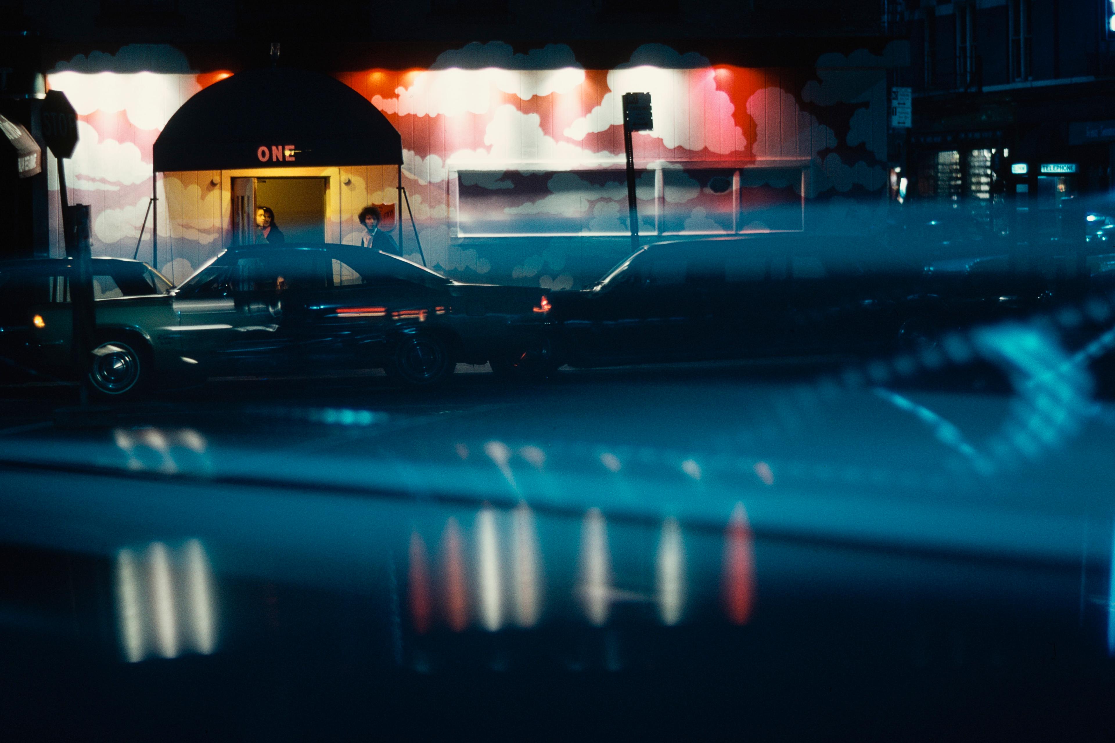 Photo of a neon-lit street scene with parked cars at night, featuring a mural with clouds on a building entrance.