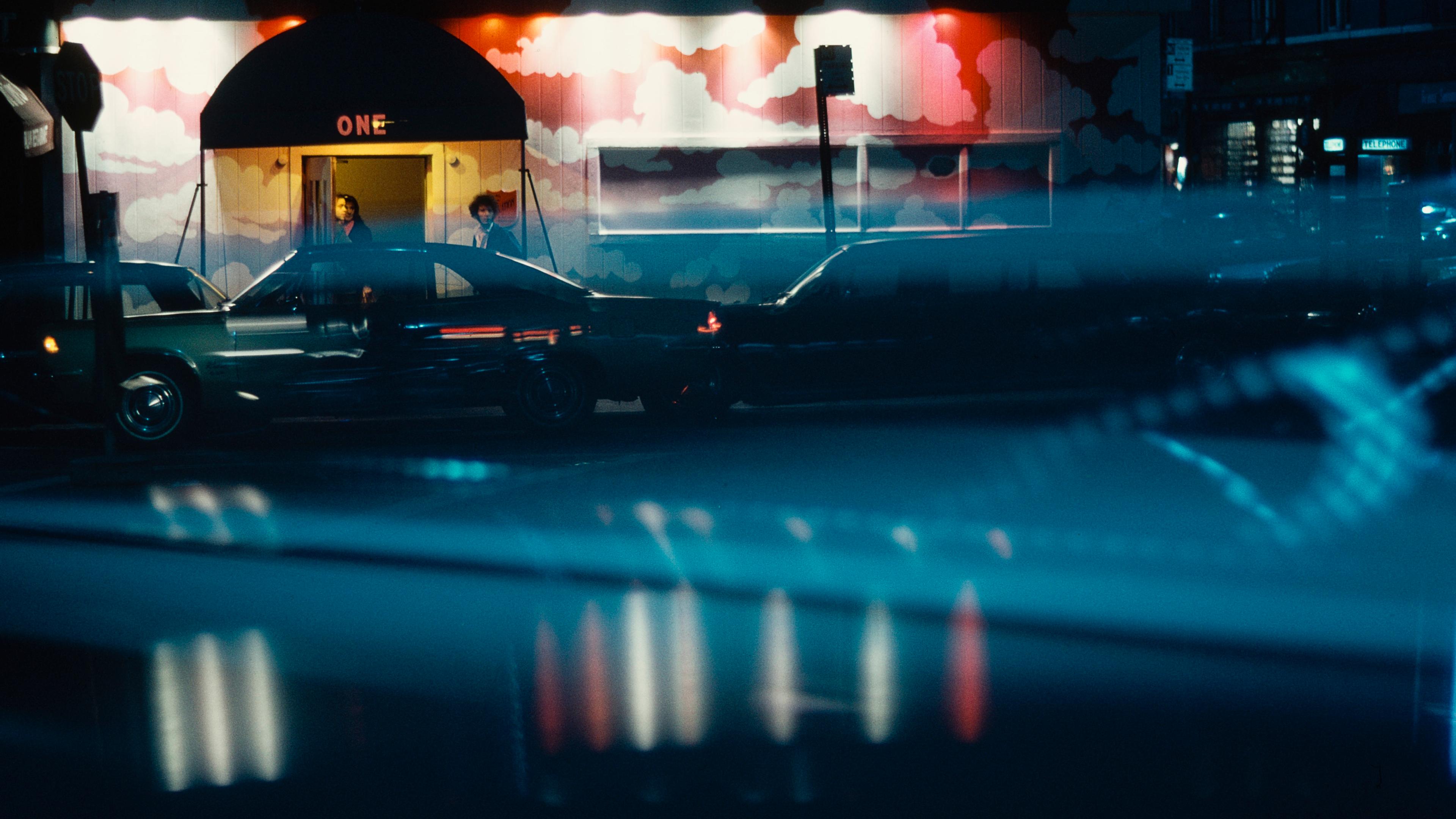 Photo of a neon-lit street scene with parked cars at night, featuring a mural with clouds on a building entrance.