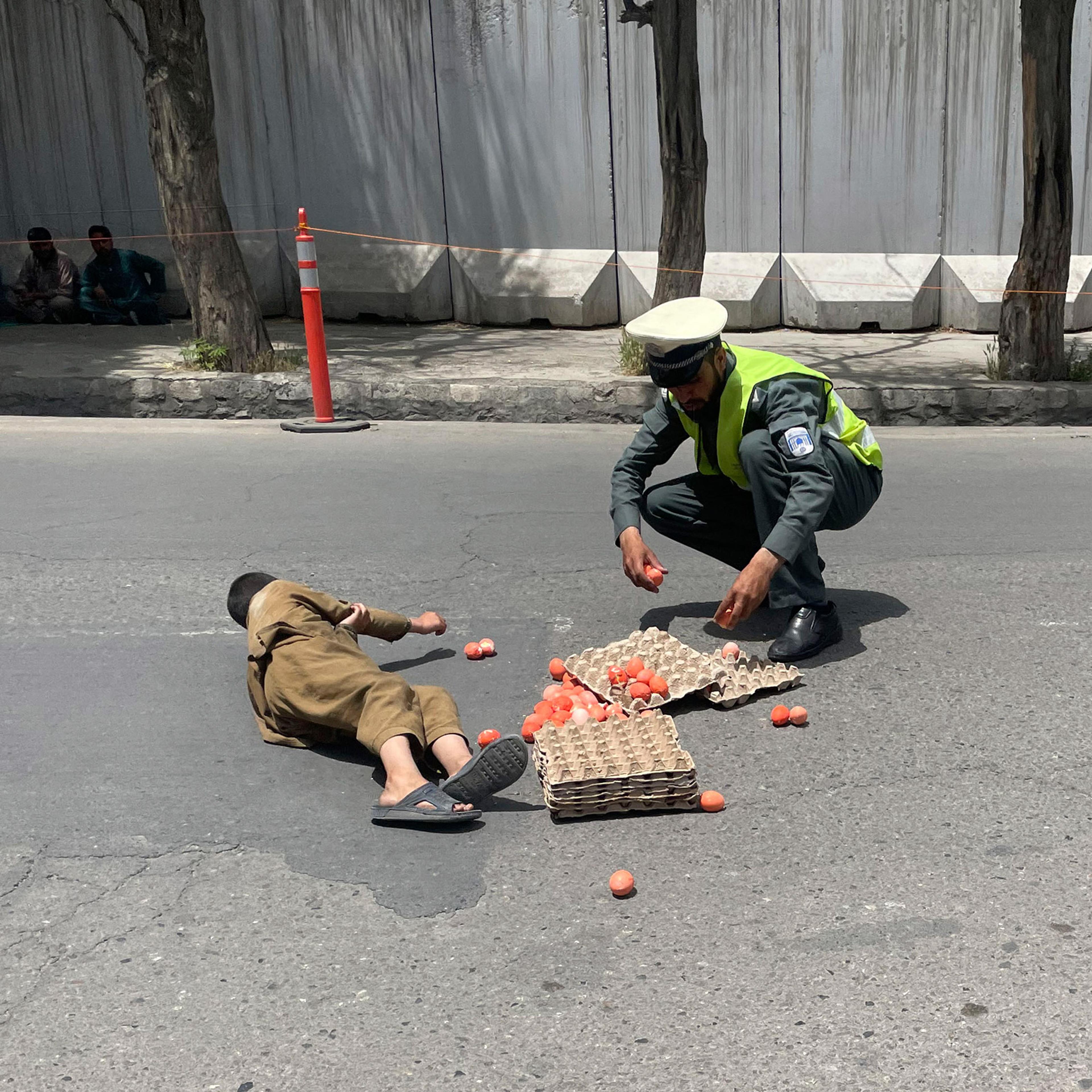 A child in traditional clothing lies on a grey street surrounded by eggs and egg trays whilst a policeman trys to collect them for him