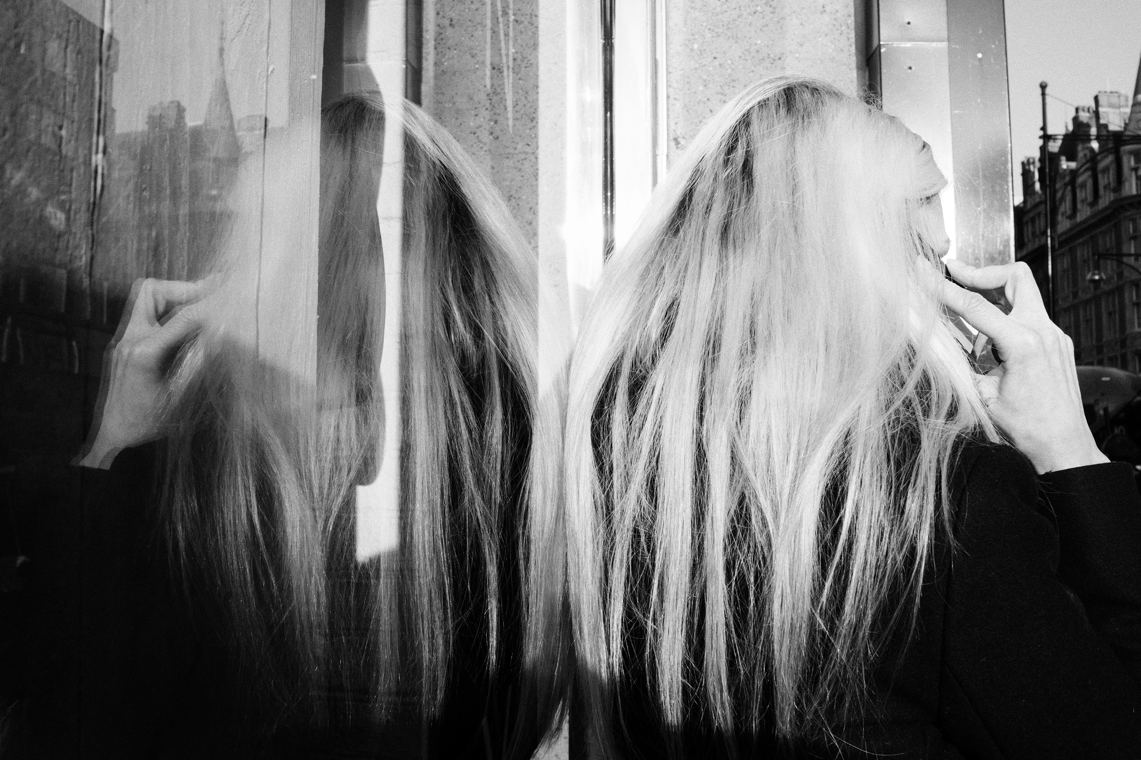 Black and white photo of a person with long hair reflected in glass talking on a mobile phone in an urban setting.