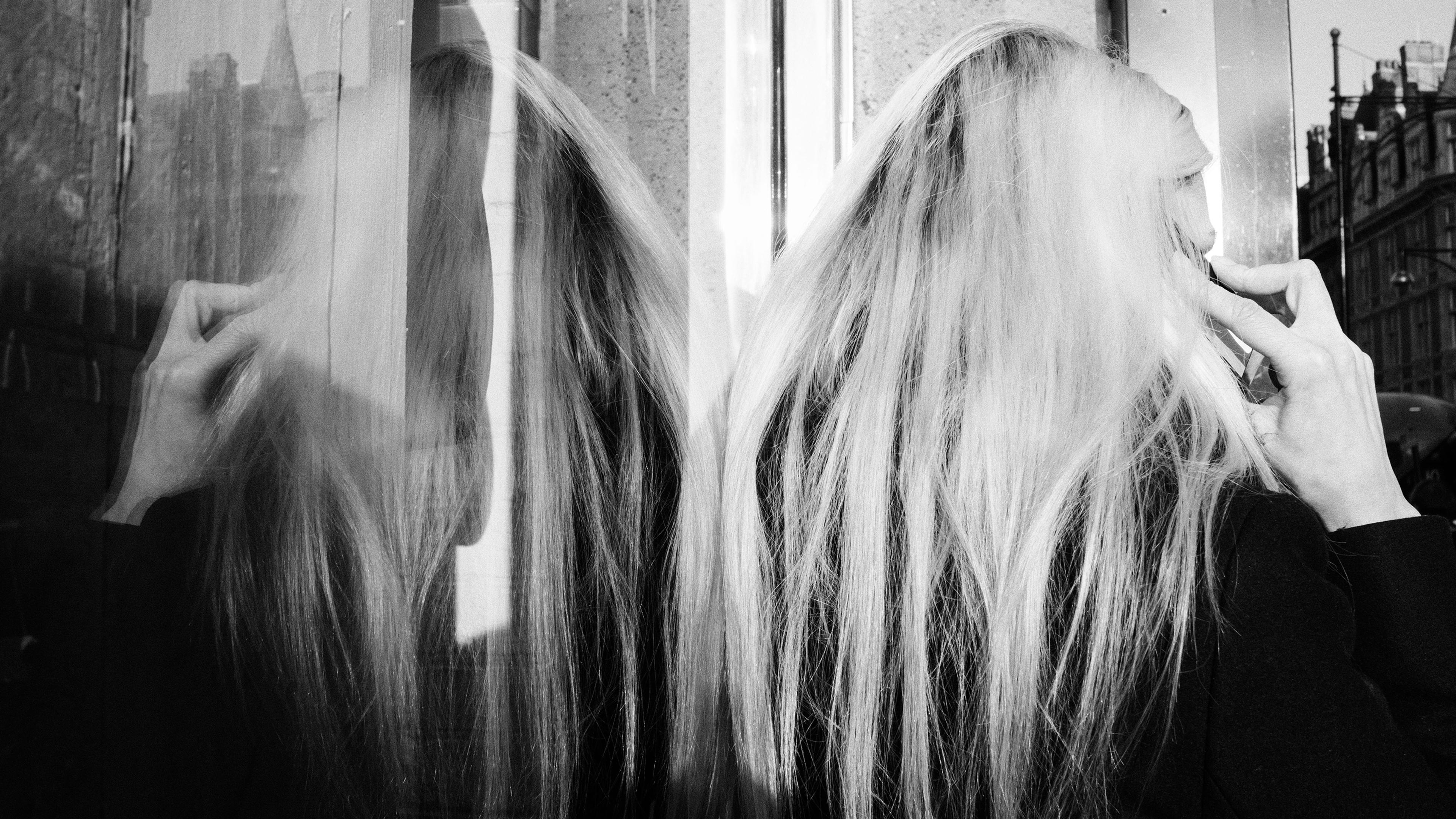Black and white photo of a person with long hair reflected in glass talking on a mobile phone in an urban setting.