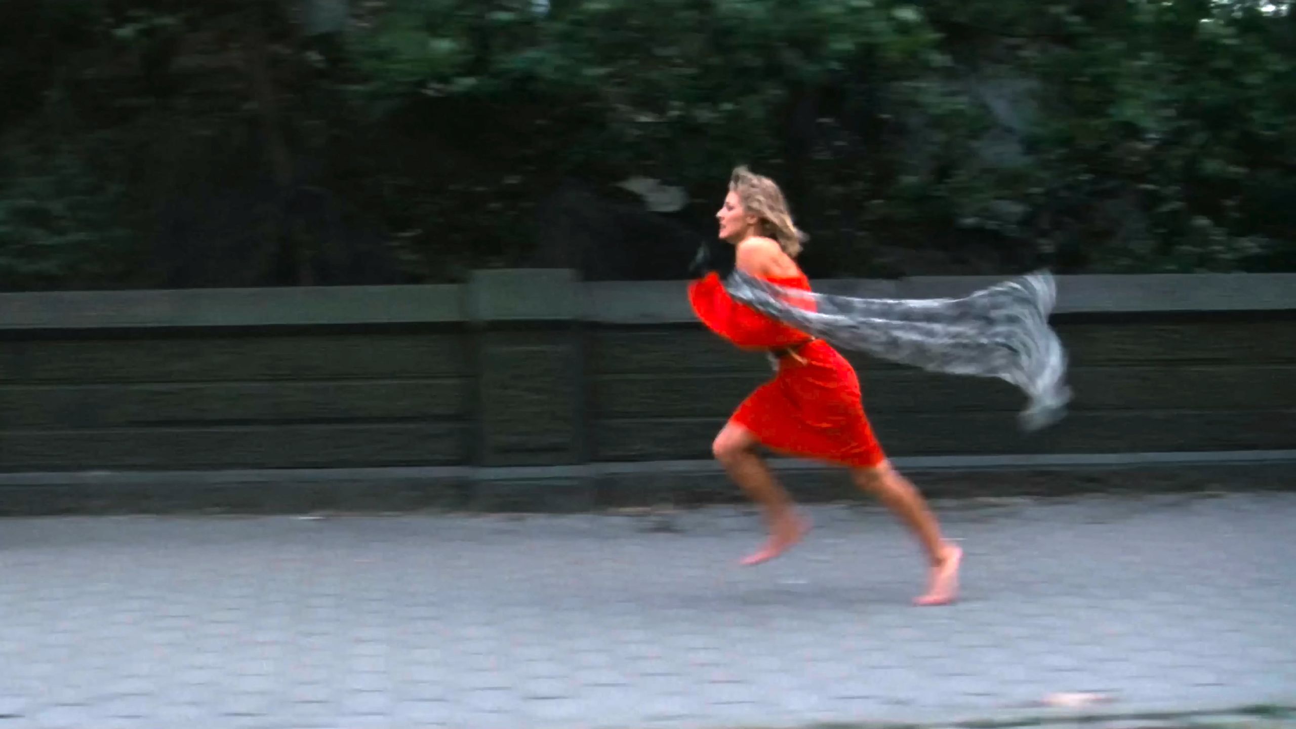 Photo of a woman in a red dress running barefoot on a path with a scarf trailing behind her, greenery in the background.