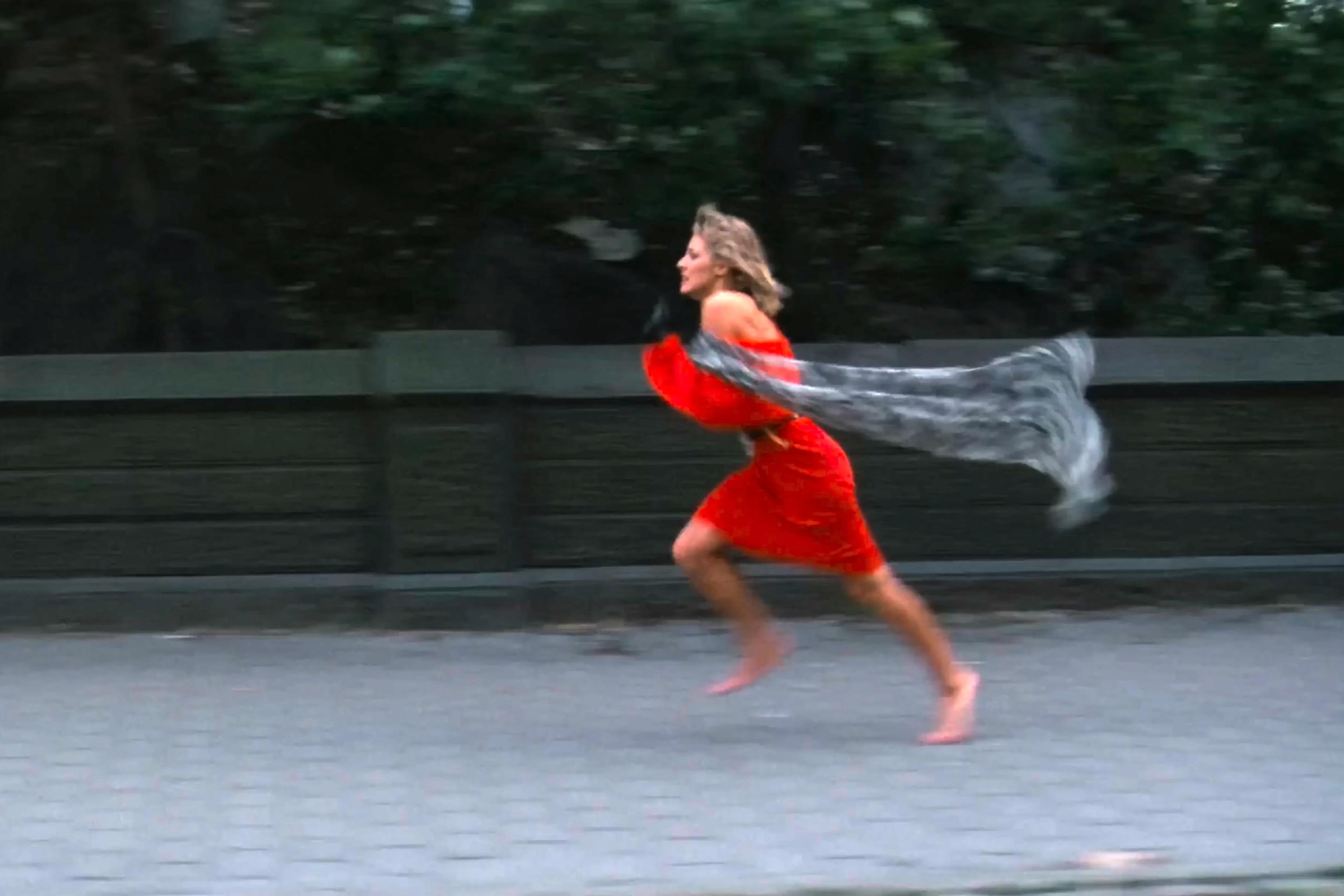 Photo of a woman in a red dress running barefoot on a path with a scarf trailing behind her, greenery in the background.