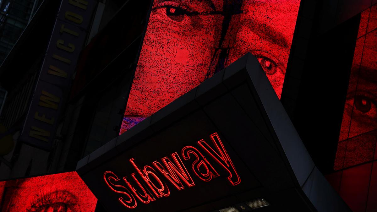 A subway sign with red neon text and large images of eyes on illuminated red panels above.