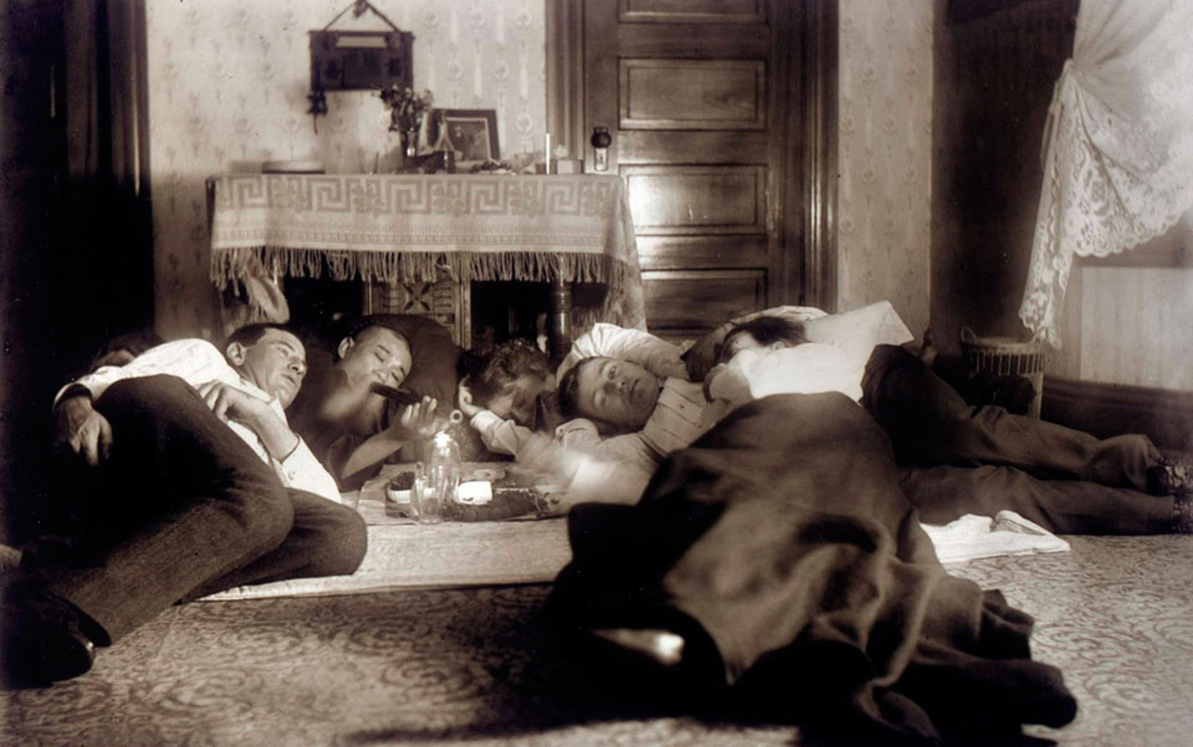 Five people lying on the floor around a lamp in a vintage-style room with a patterned carpet and a wooden door.