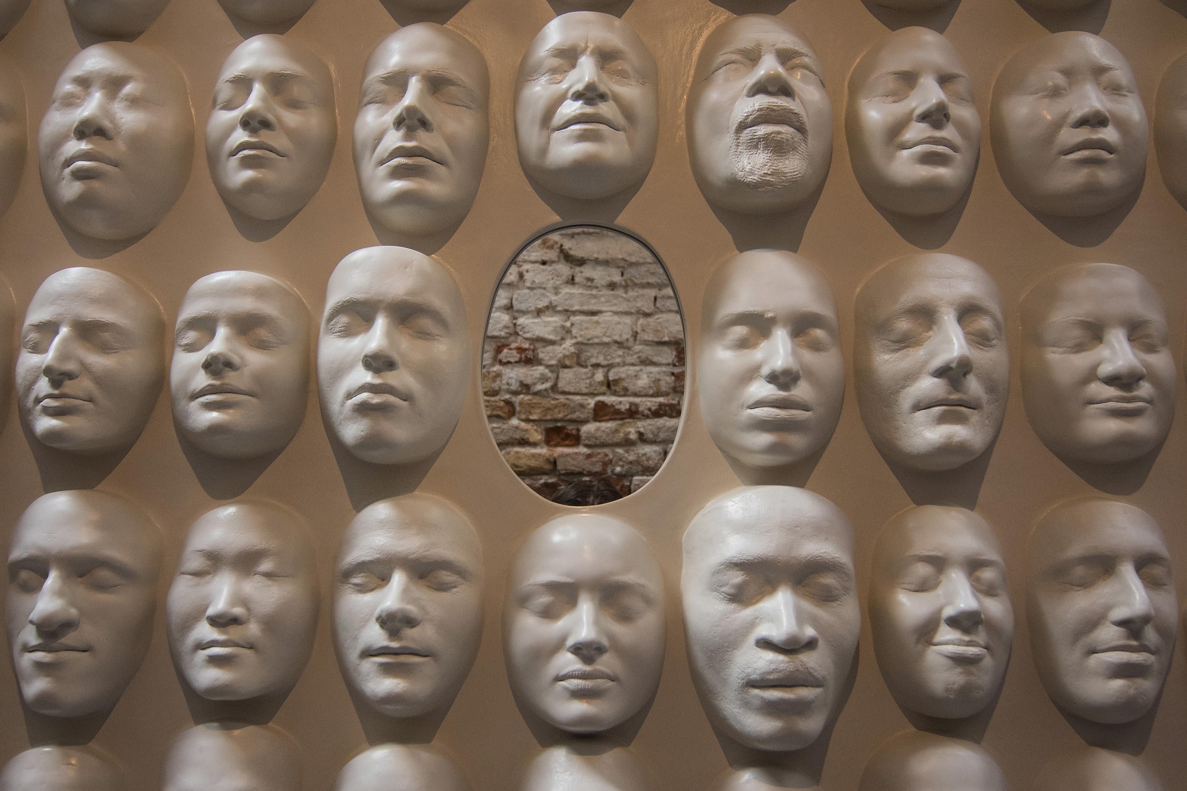 Wall sculpture with white human face masks surrounding an oval gap revealing a brick wall behind.
