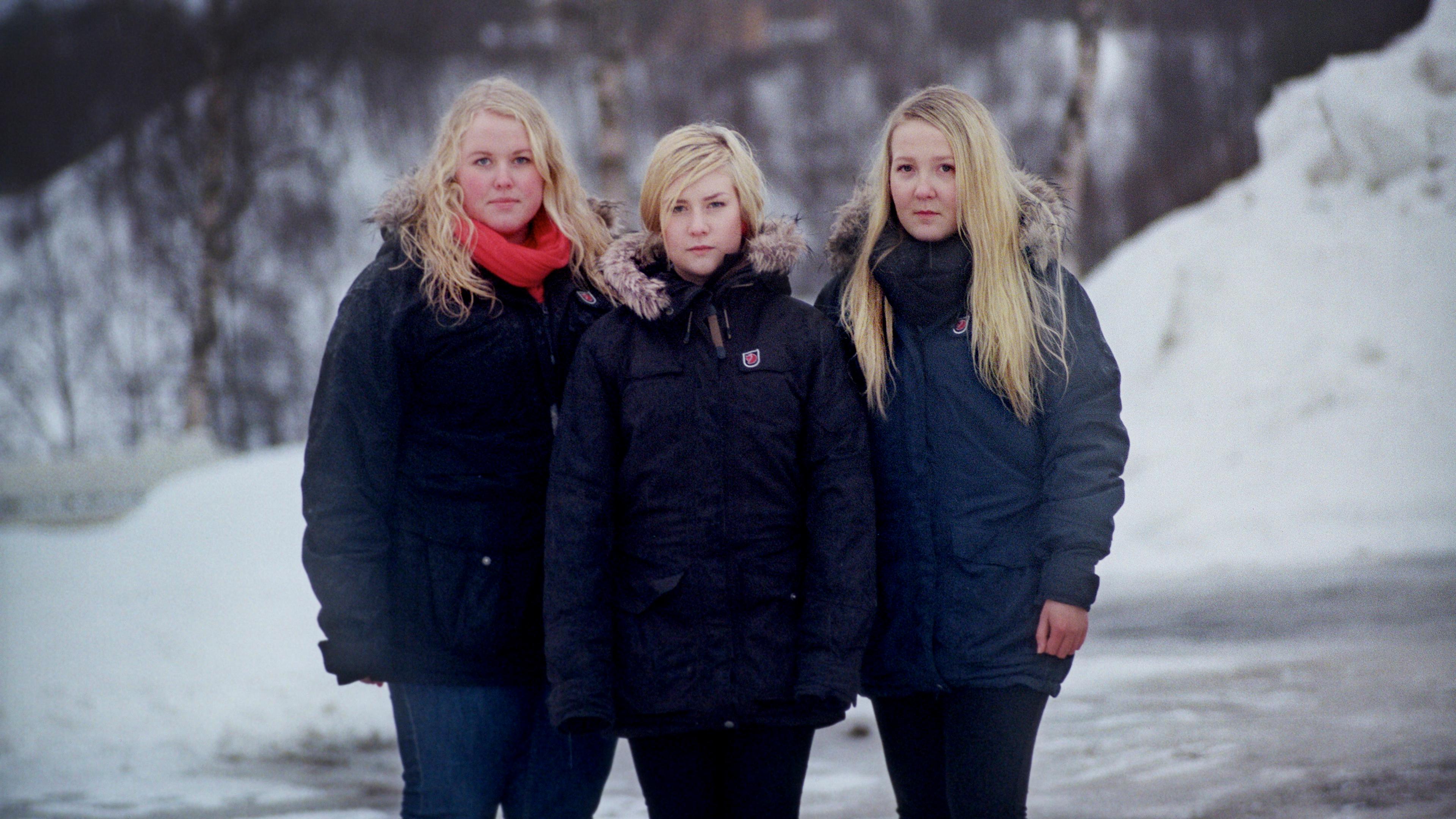 Photo of three people in winter coats standing on a snowy path with bare trees in the background.