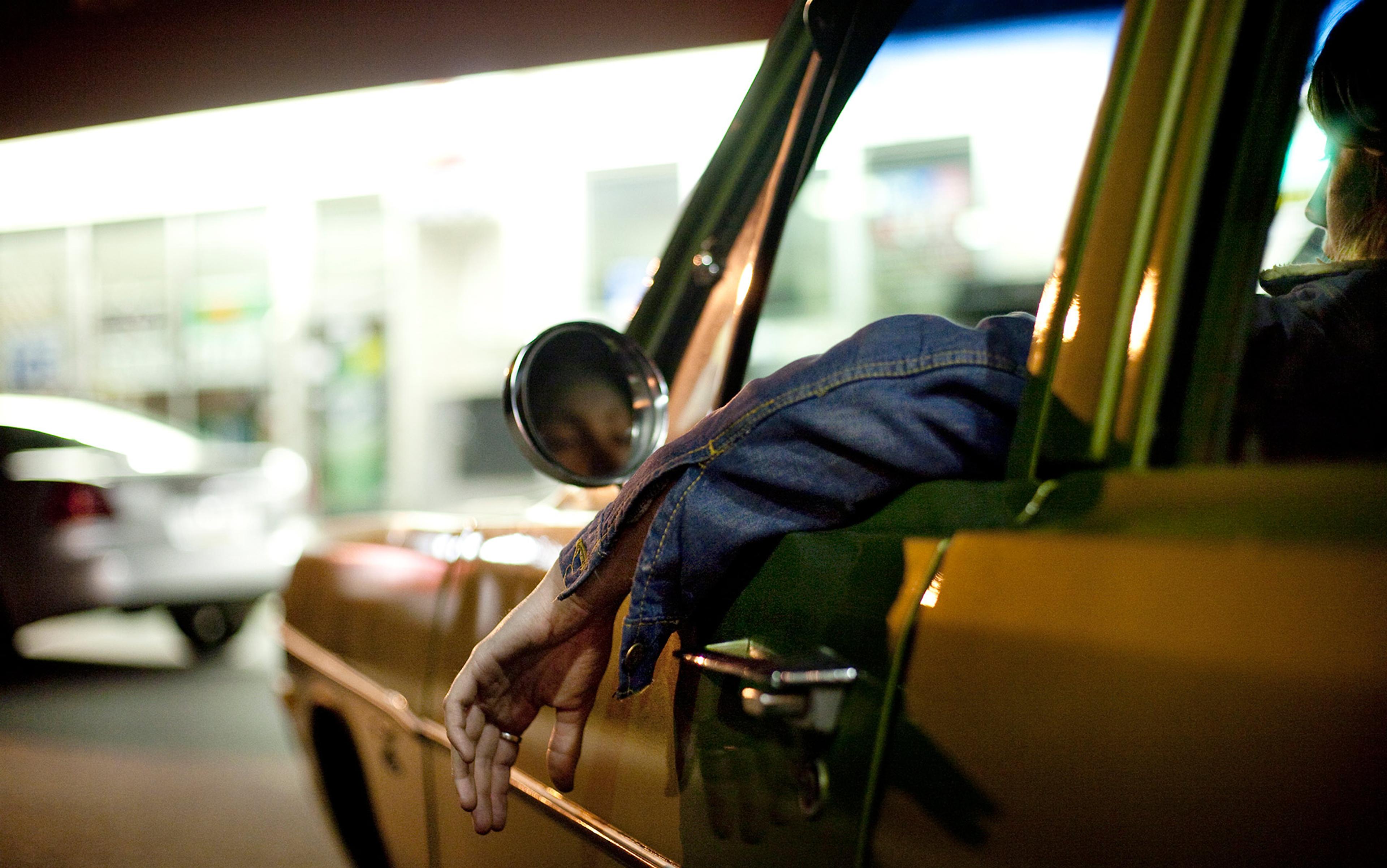 A person in a yellow car at night, with their arm draped out of the window. The background shows blurred lights and a building.