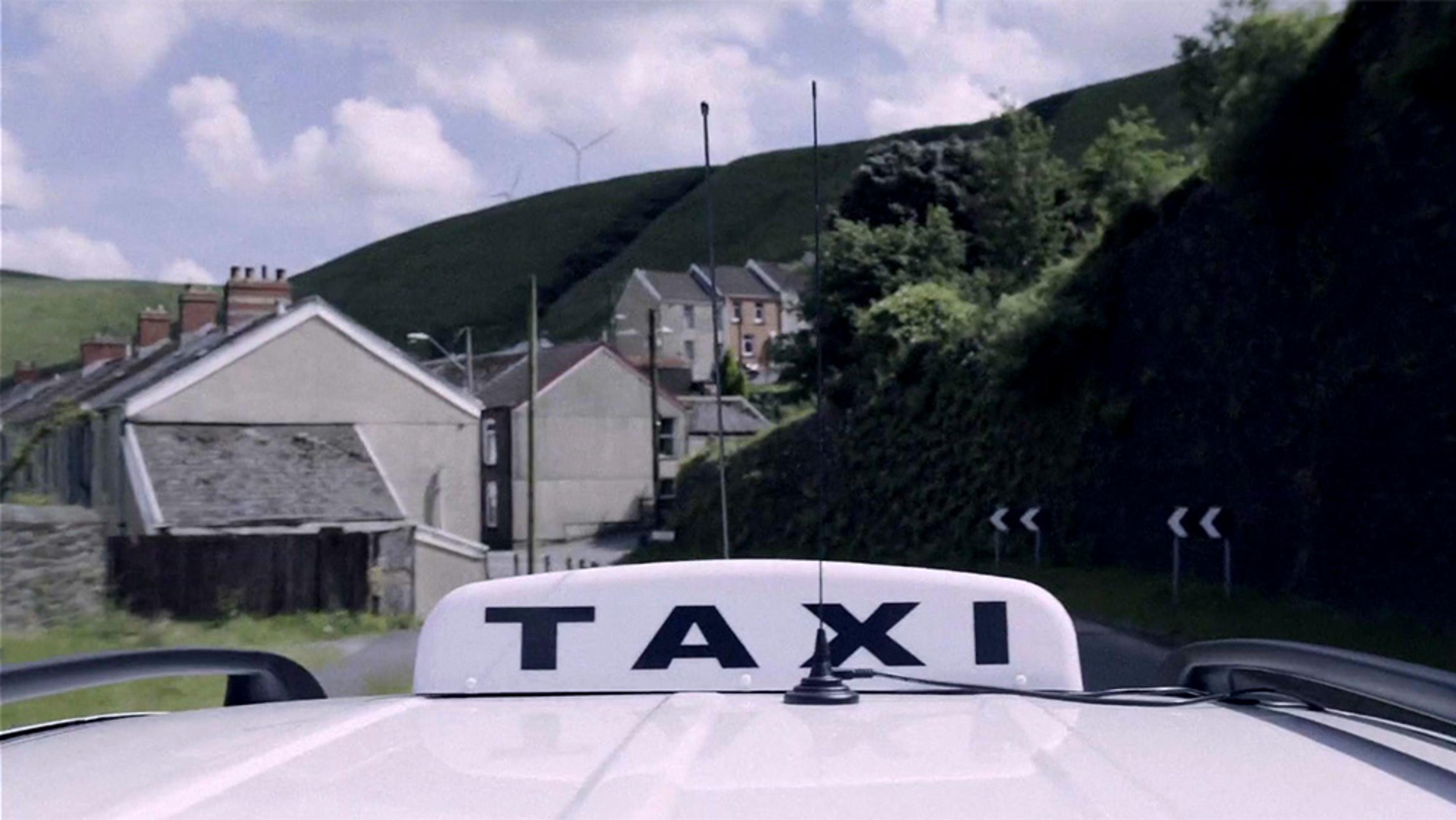 A taxi roof sign with houses and green hills in the background, under a partly cloudy sky in a rural area.