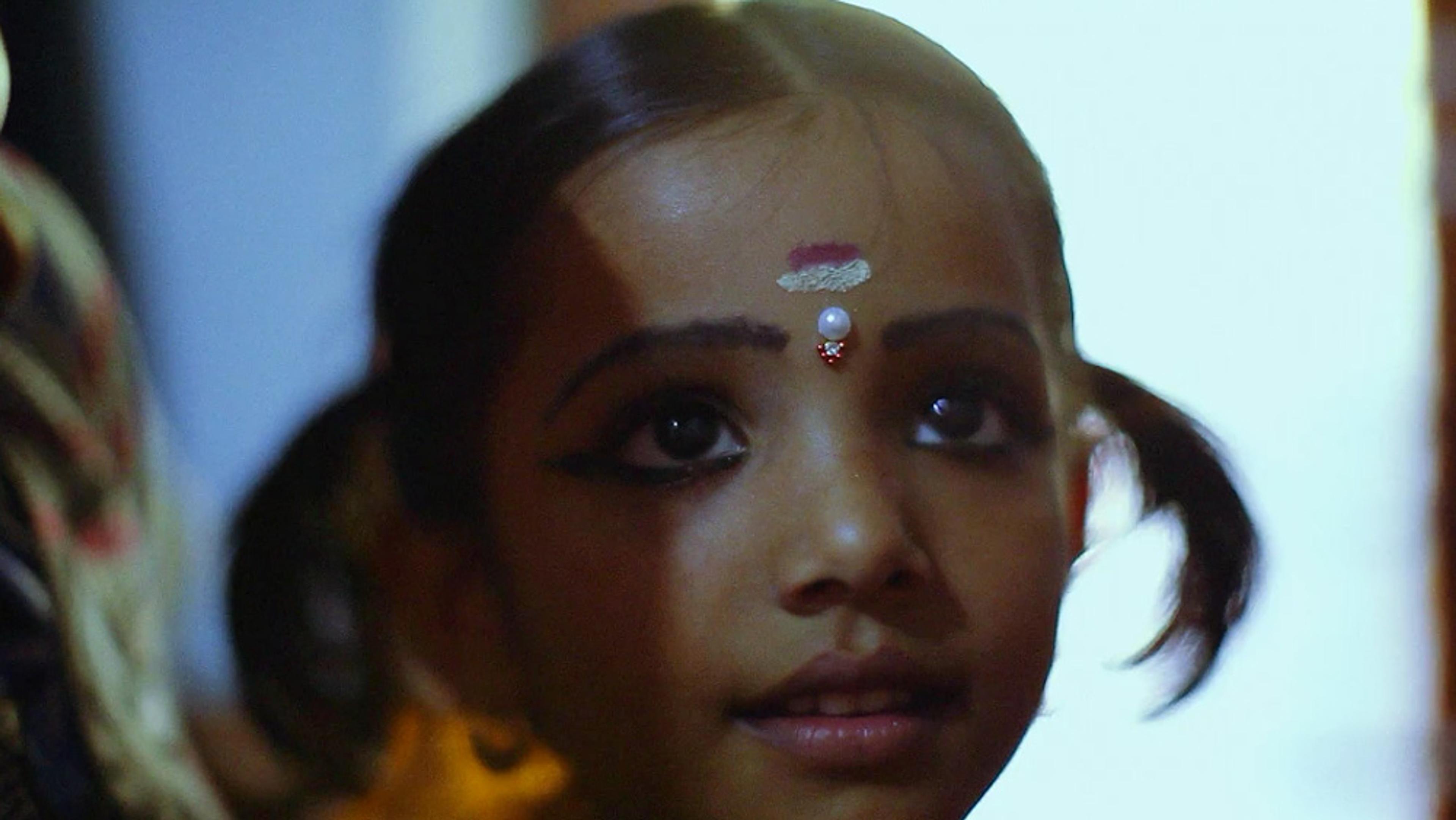 Close-up of a child with facial decorations and pigtails, looking upwards, illuminated softly against a blurred background.