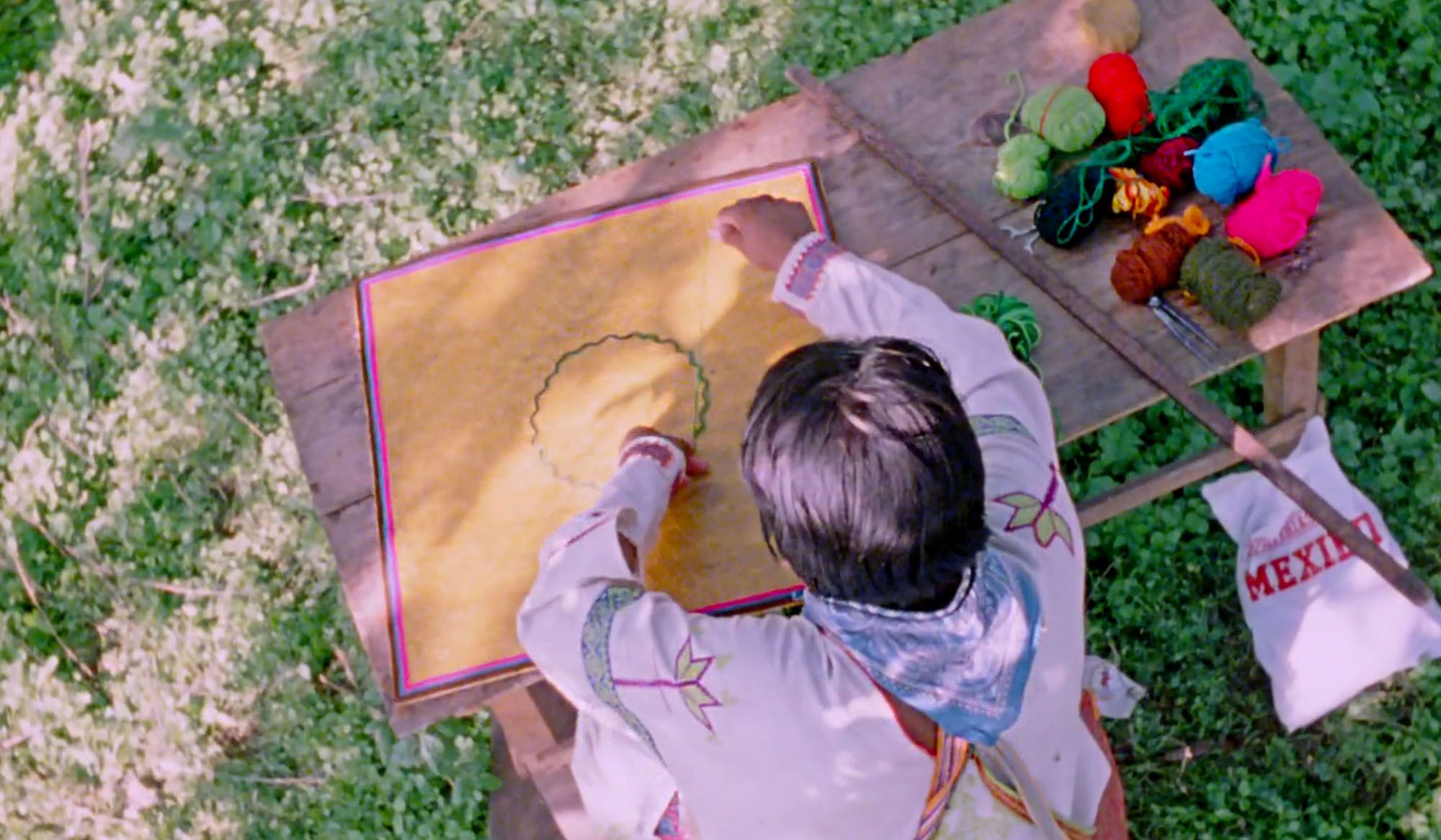 A person in traditional clothing embroiders a yellow fabric on a table outdoors, with assorted yarn and tools beside them.