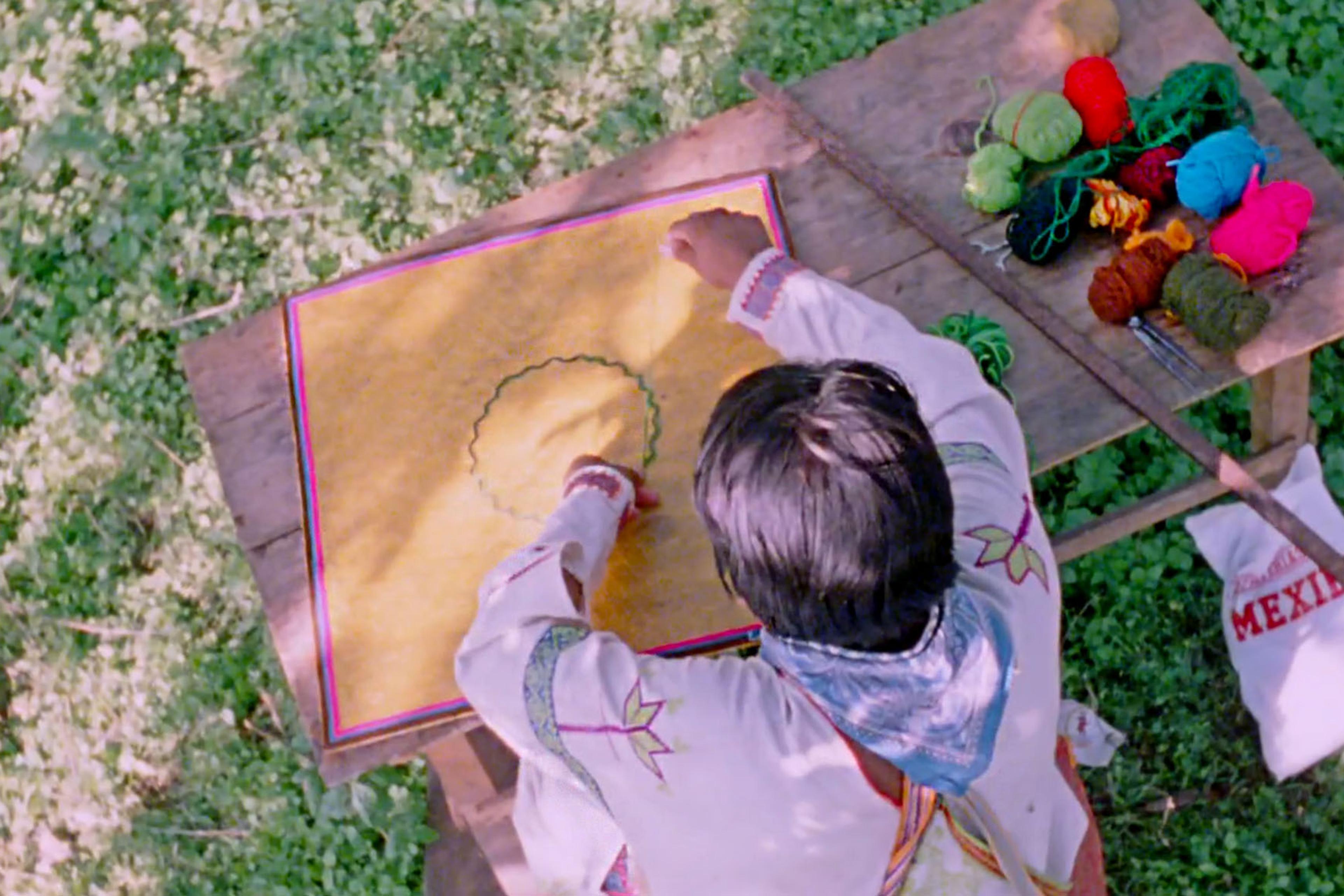 A person in traditional clothing embroiders a yellow fabric on a table outdoors, with assorted yarn and tools beside them.