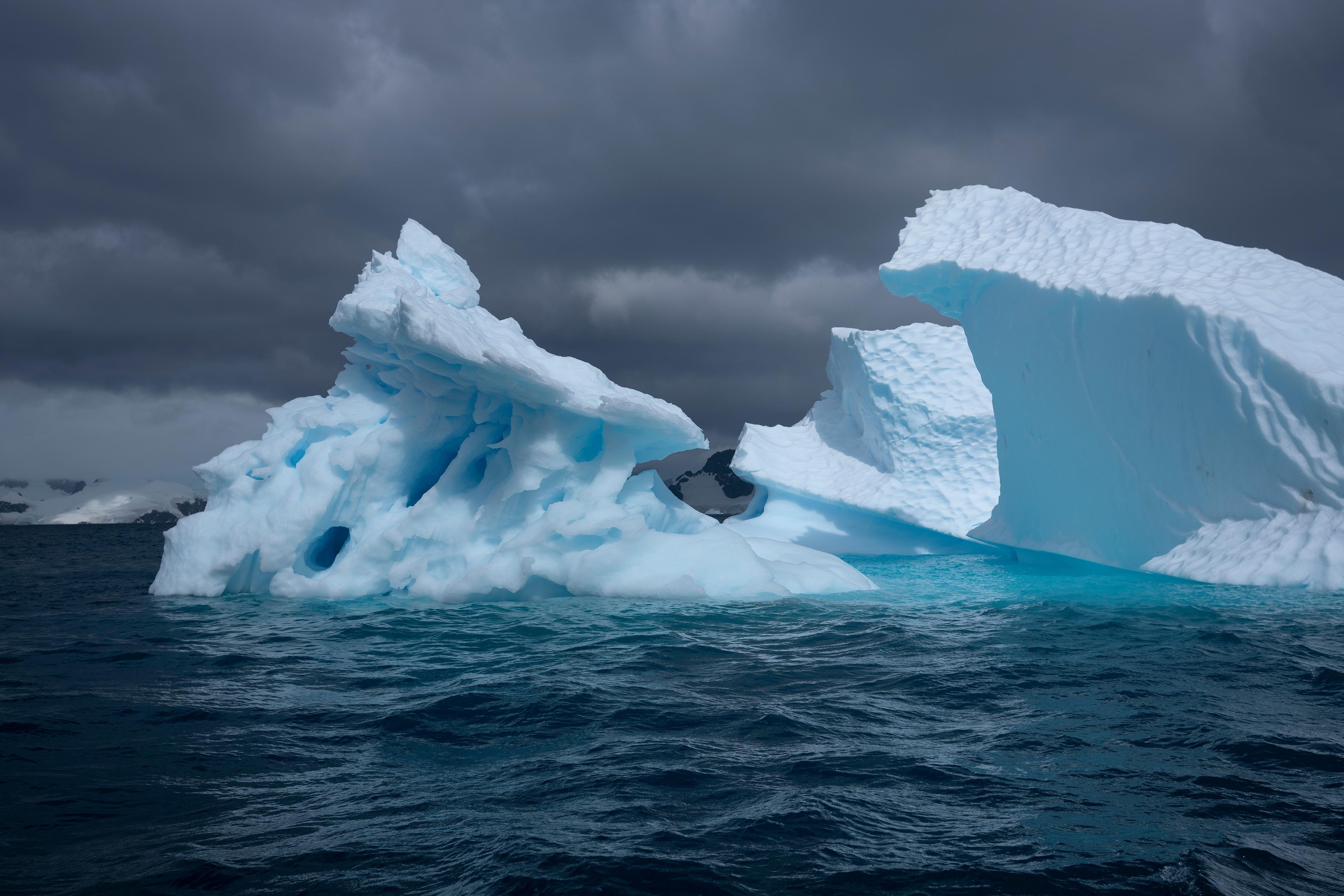 Photo of large jagged icebergs in dark ocean waters under a cloudy sky.