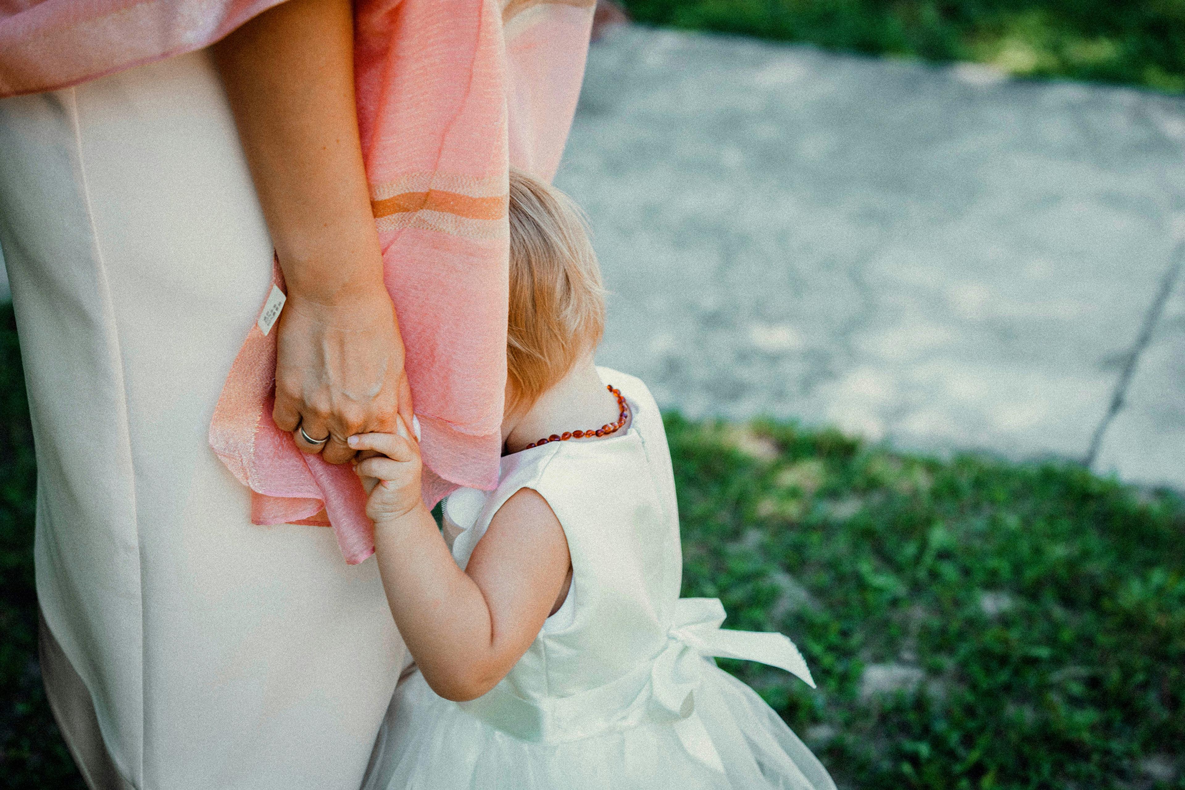 A child in a white dress clinging to a woman’s hand, hiding her face in the woman’s pink shawl, outdoors.