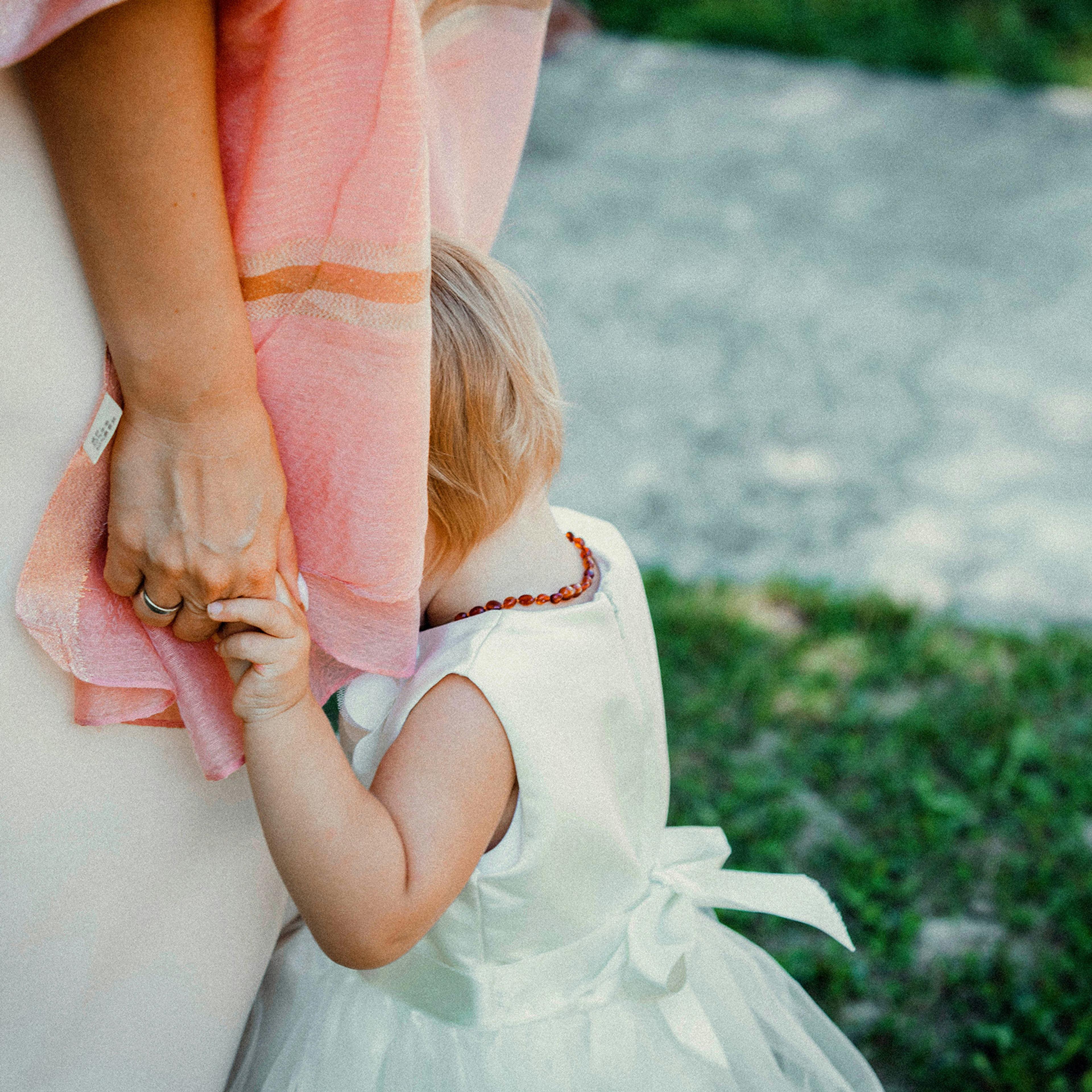 A child in a white dress clinging to a woman’s hand, hiding her face in the woman’s pink shawl, outdoors.