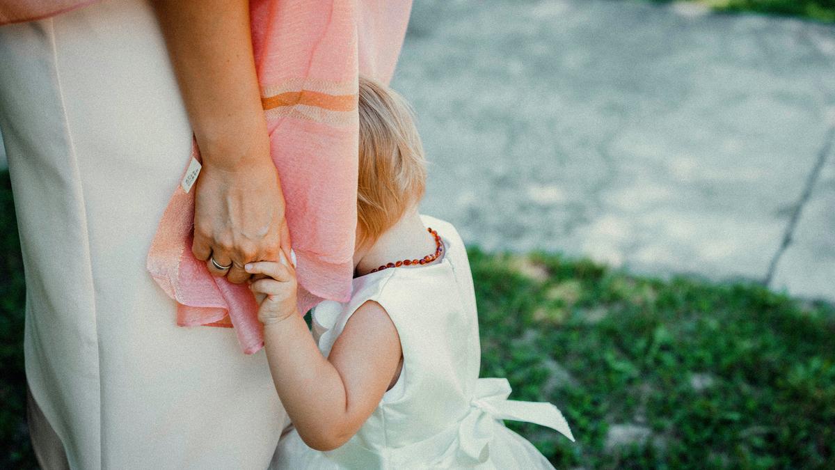 A child in a white dress clinging to a woman’s hand, hiding her face in the woman’s pink shawl, outdoors.