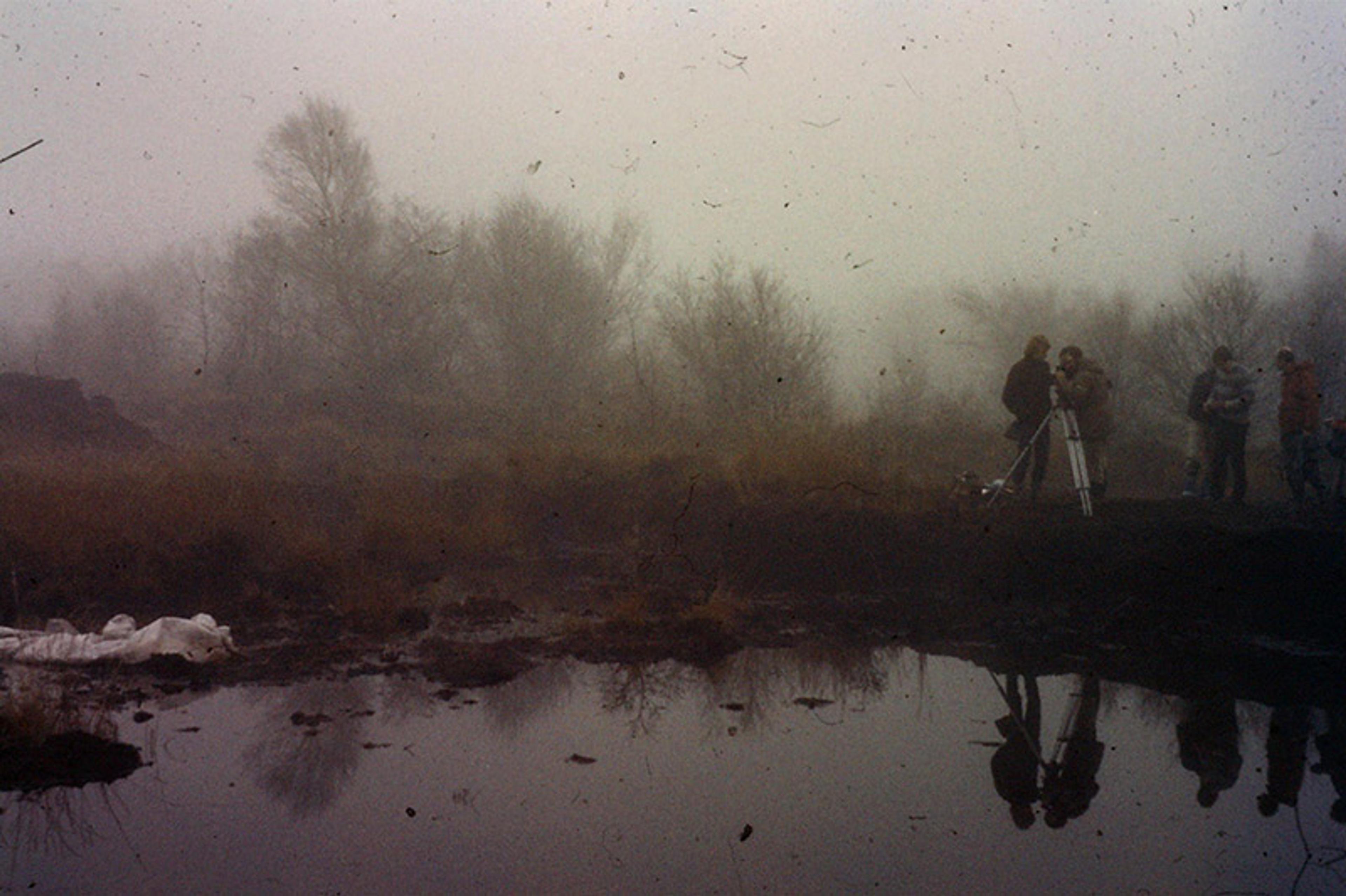 A foggy landscape photo with trees, a pond and several people gathered around a camera setup on a tripod.