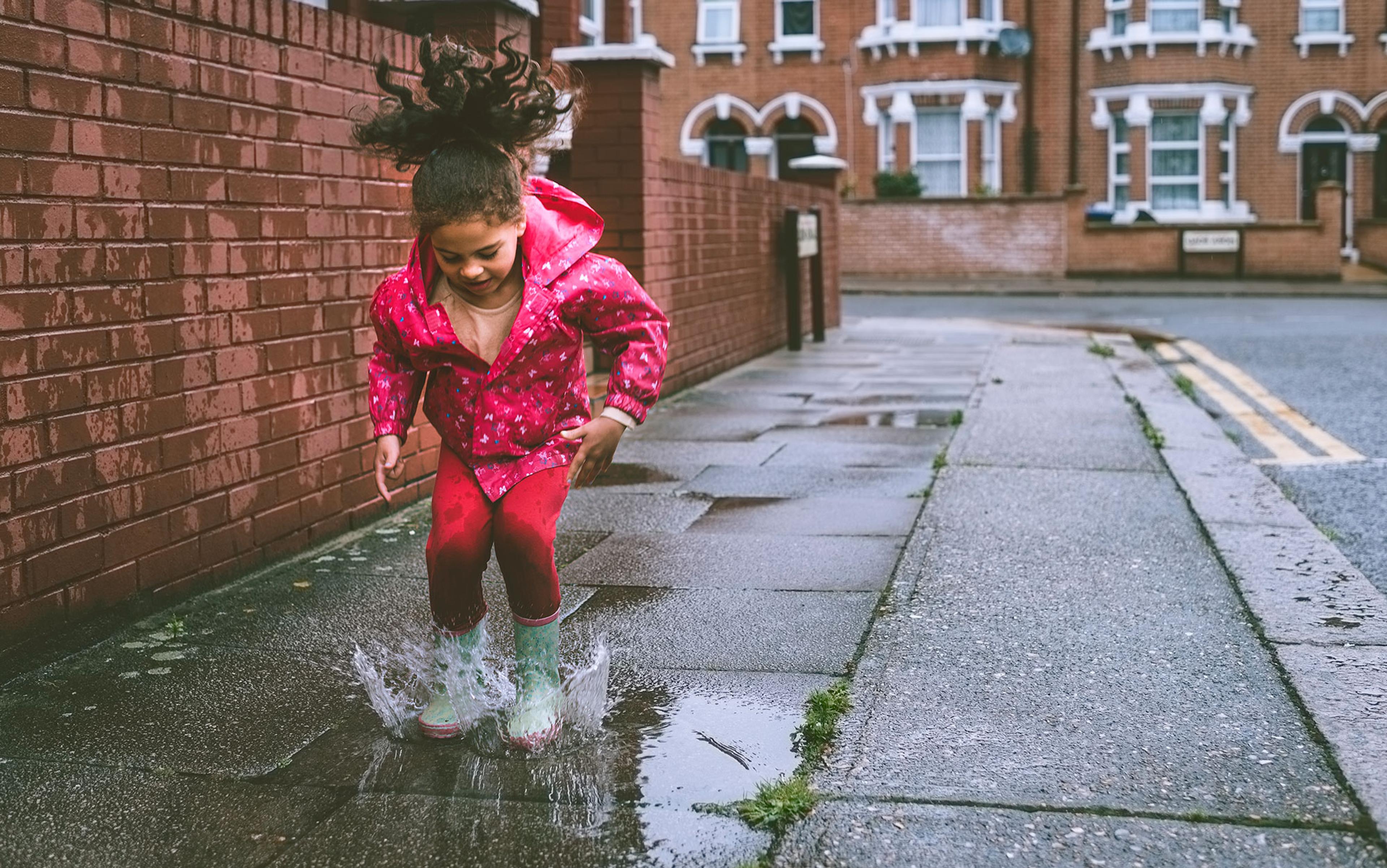 A child in a pink raincoat jumping in a puddle on a city street on a rainy day with terraced houses in the background.