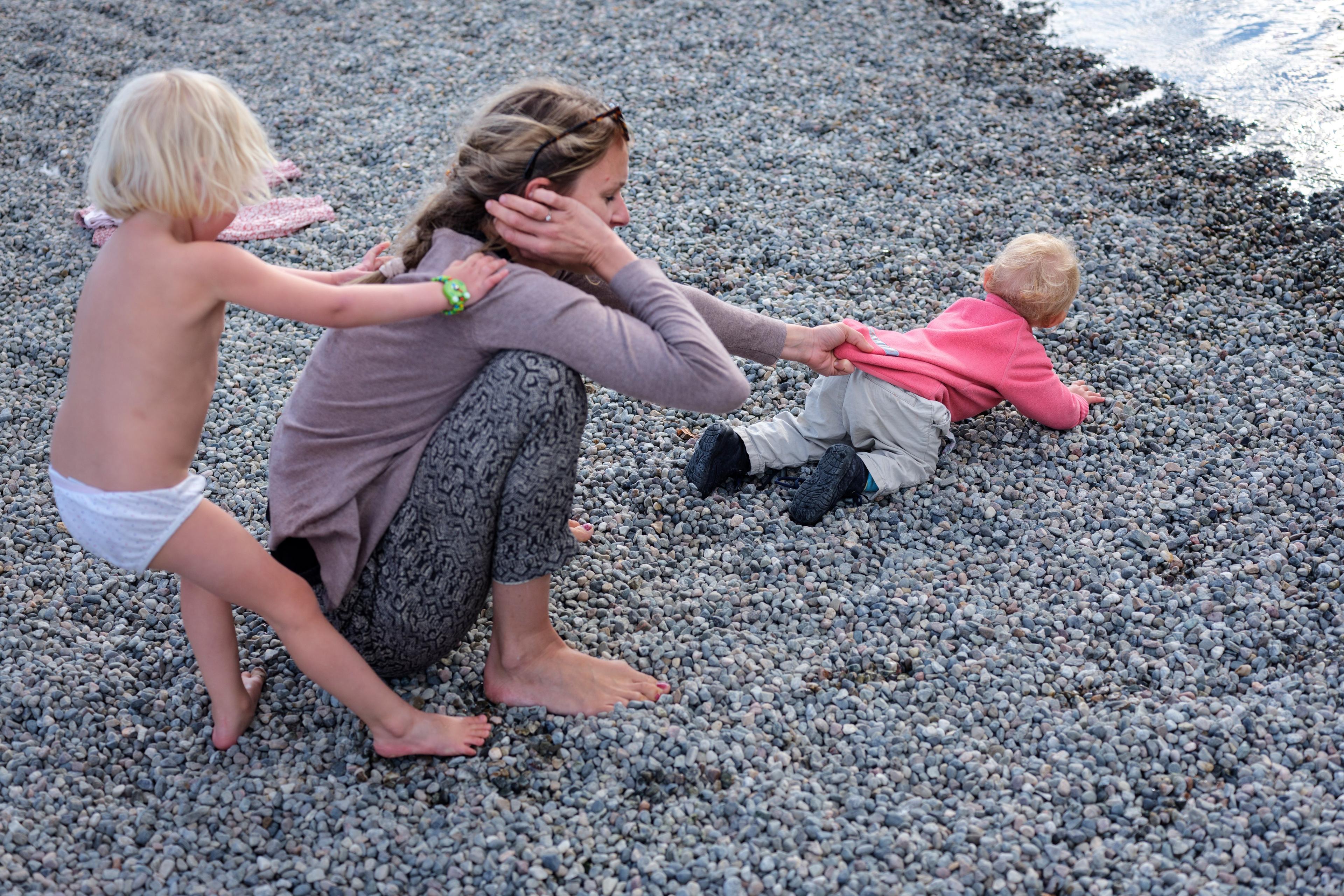 Photo of a woman on a pebble beach with two children, one pulling her hair and the other crawling towards the water.