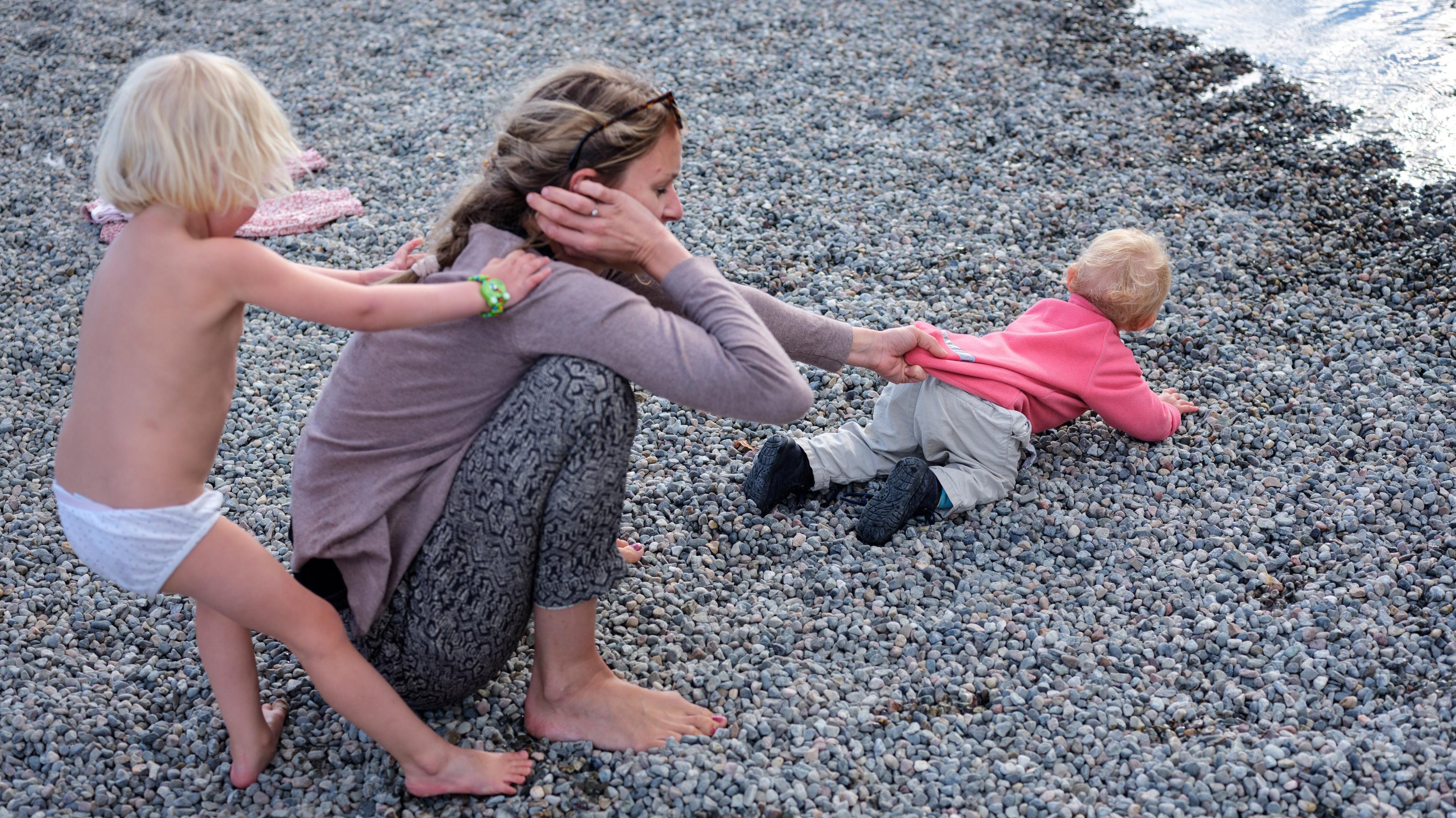 Photo of a woman on a pebble beach with two children, one pulling her hair and the other crawling towards the water.