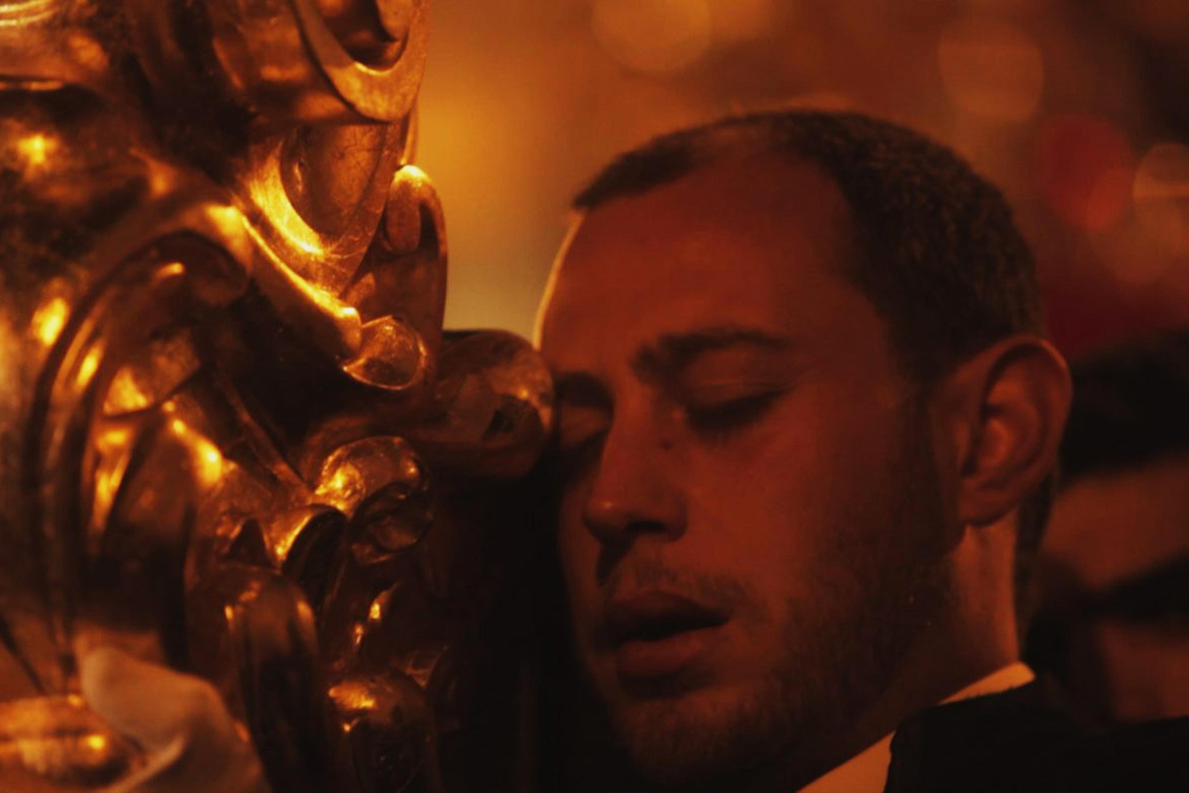 A man with his eyes closed leaning his head against a golden object in a dimly lit setting.