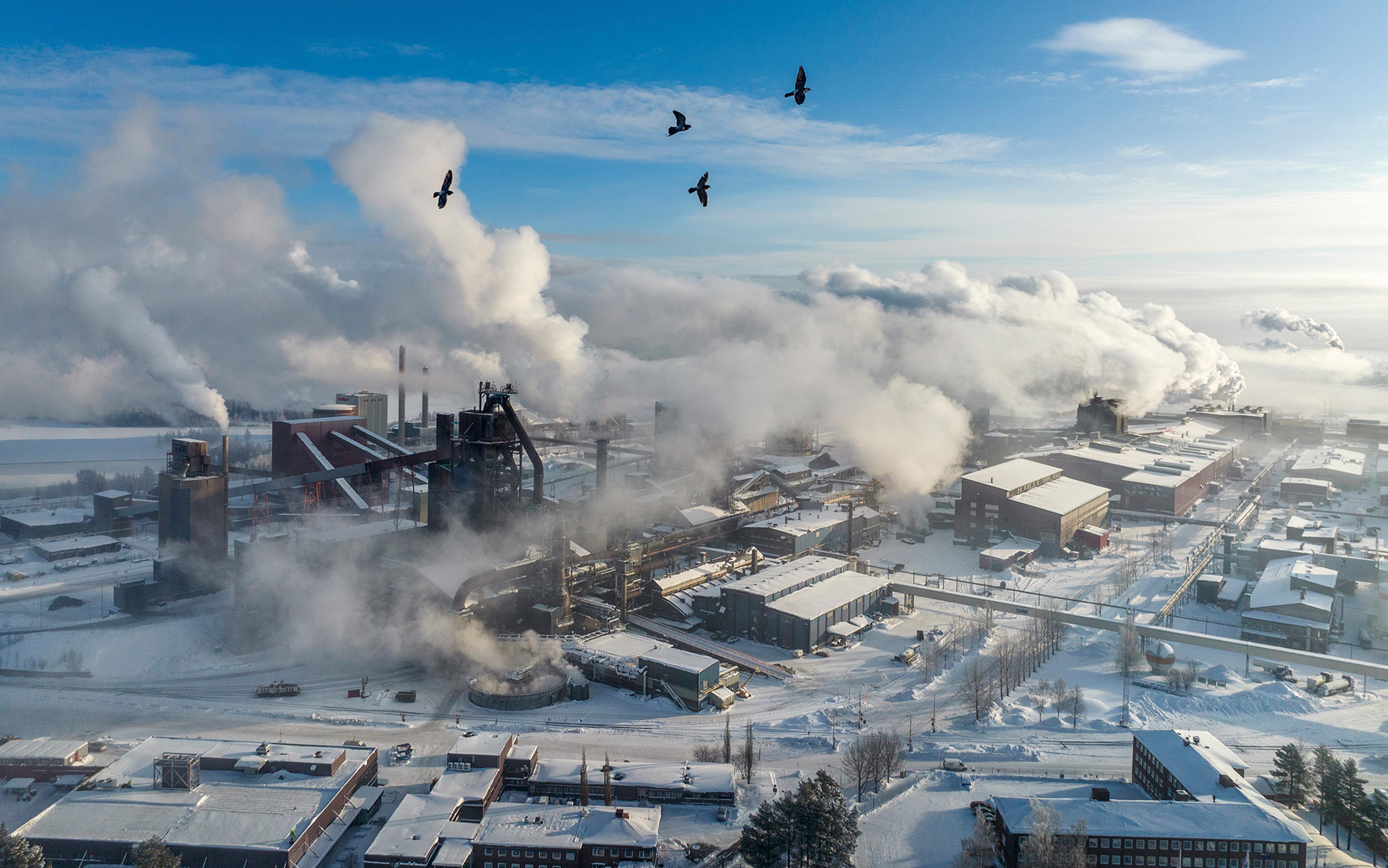 Aerial view of an industrial site emitting smoke, surrounded by snow-covered buildings and landscape, under a clear blue sky with birds flying overhead.