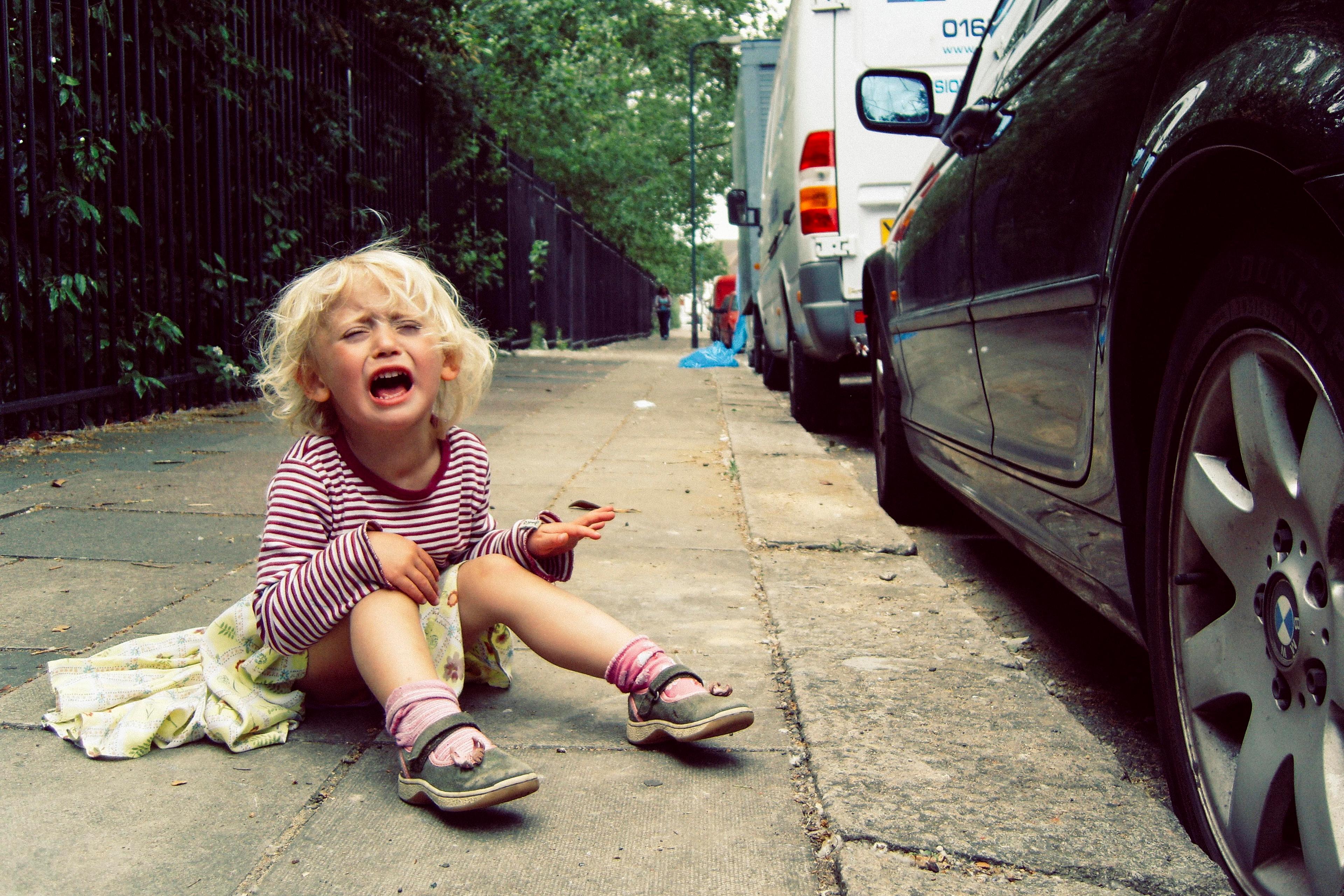 Photo of a child in a striped shirt sitting on a pavement crying, with cars parked along the street in the background.